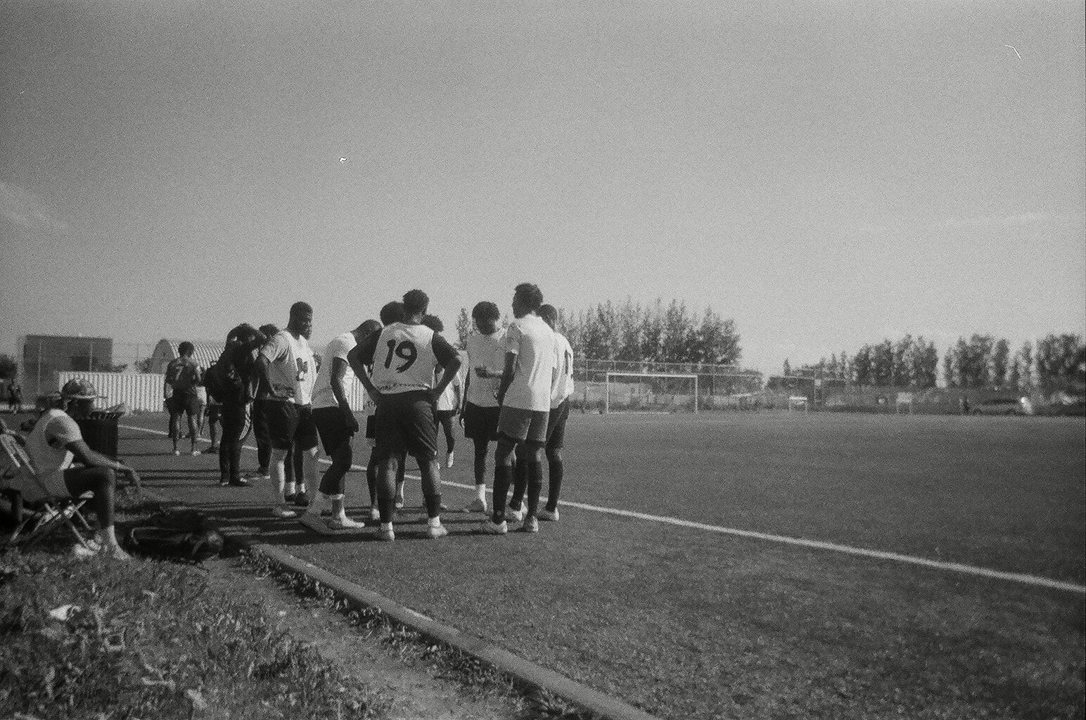 A group of soccer players in uniforms standing together on a soccer field, with some sitting on benches, under a clear sky.