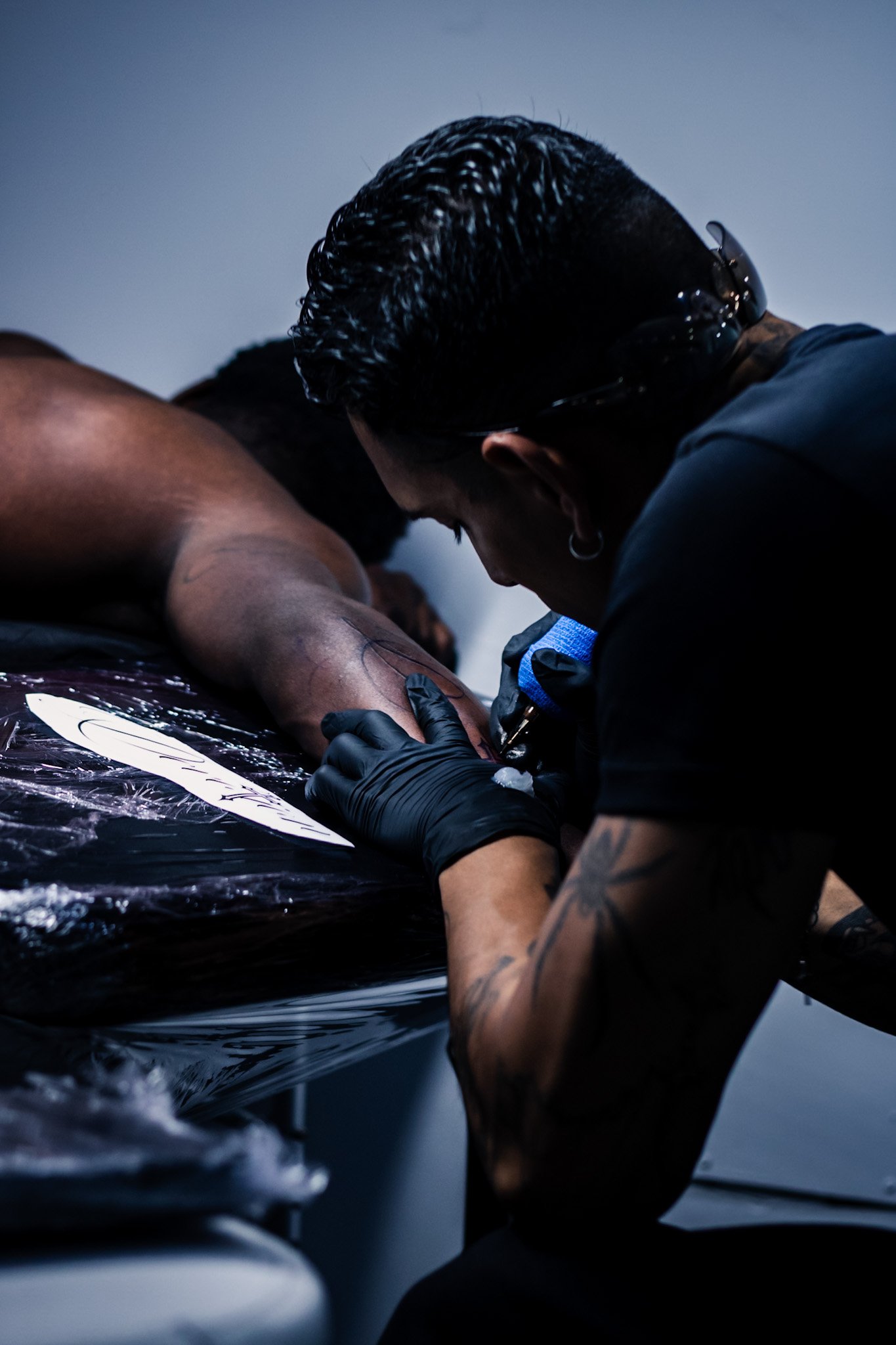 A tattooed person with glasses is tattooing another person's arm in a tattoo studio. The person receiving the tattoo is lying down on a surface covered with clear plastic.
