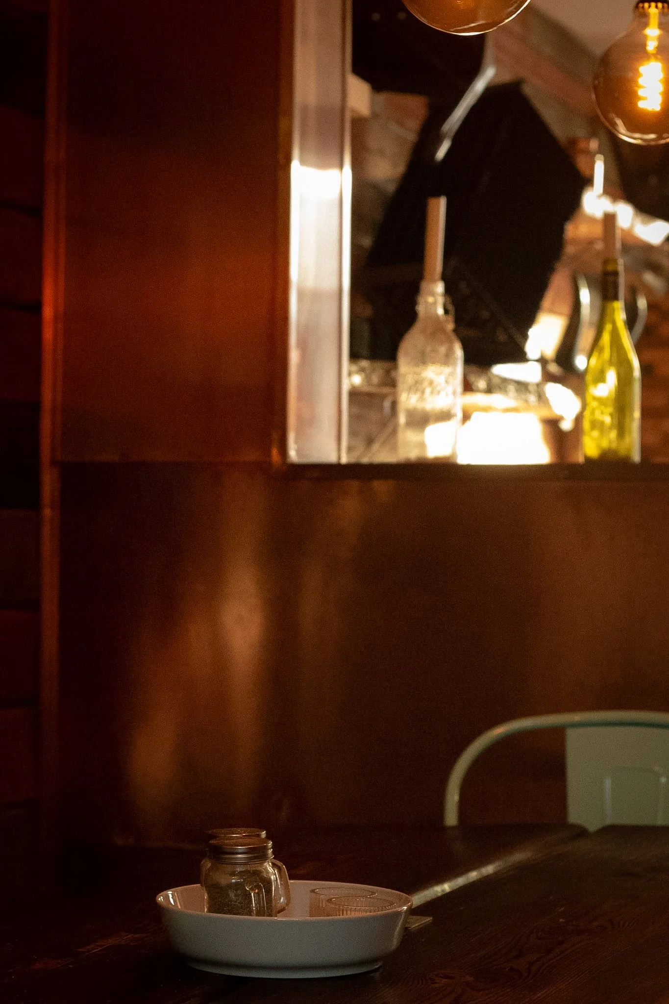 Cozy restaurant or café setting with dim lighting, a small glass jar, a white bowl and a paper plate on a dark wooden table, and warm hanging lights in the background.
