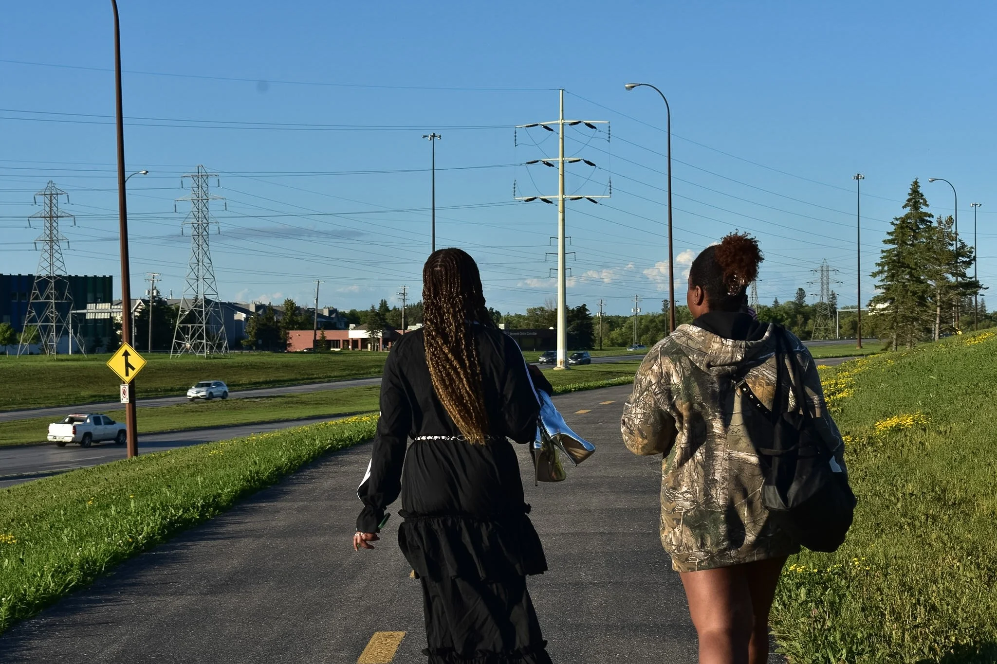 Two women walking along a paved path beside a grassy area with yellow flowers, with a highway and power lines in the background.