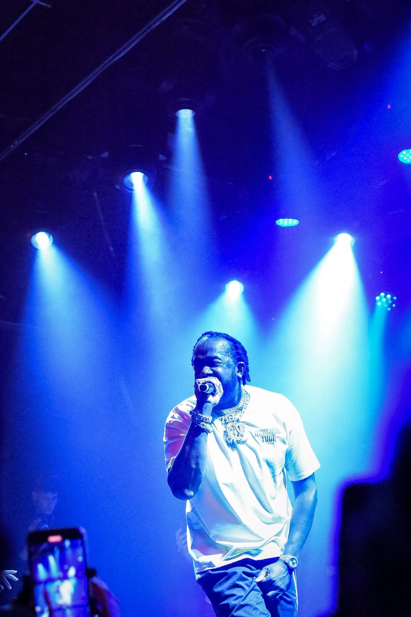 A male performer singing into a microphone under blue stage lighting, wearing a white t-shirt and jewelry.