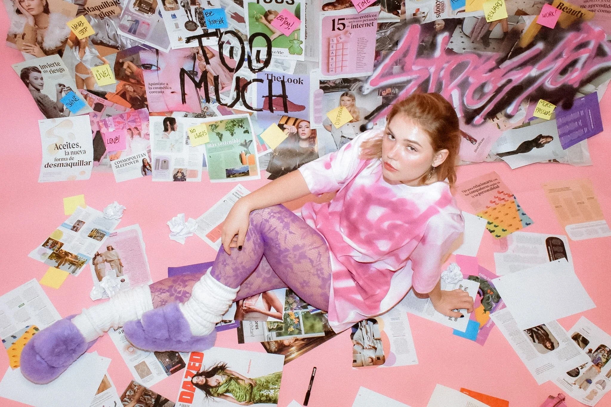 A woman with red hair lying on a pink background surrounded by torn magazine pages and sticky notes in various colors with handwritten words, with a person in purple fuzzy slippers reaching towards her.