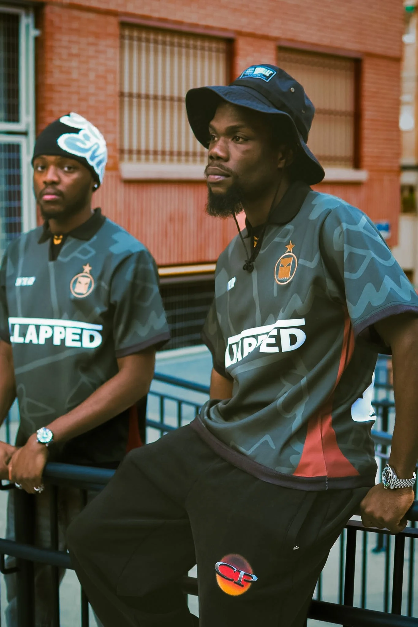 Two young men wearing skateboarding shirts with logos, posing outdoors in front of a brick building with barred windows, one wearing a bucket hat, the other a beanie, both leaning on a black railing.