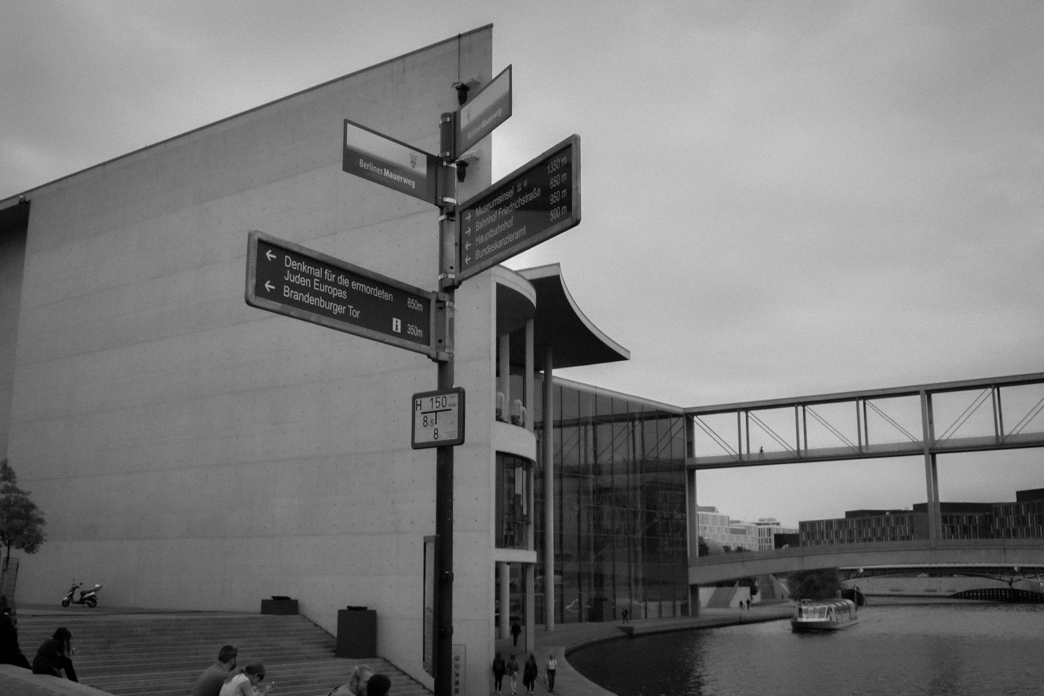 Street scene near modern buildings with multiple directional signs in German, a group of people walking, and a scooter in the foreground. There is a bridge or walkway overhead and a body of water on the right.