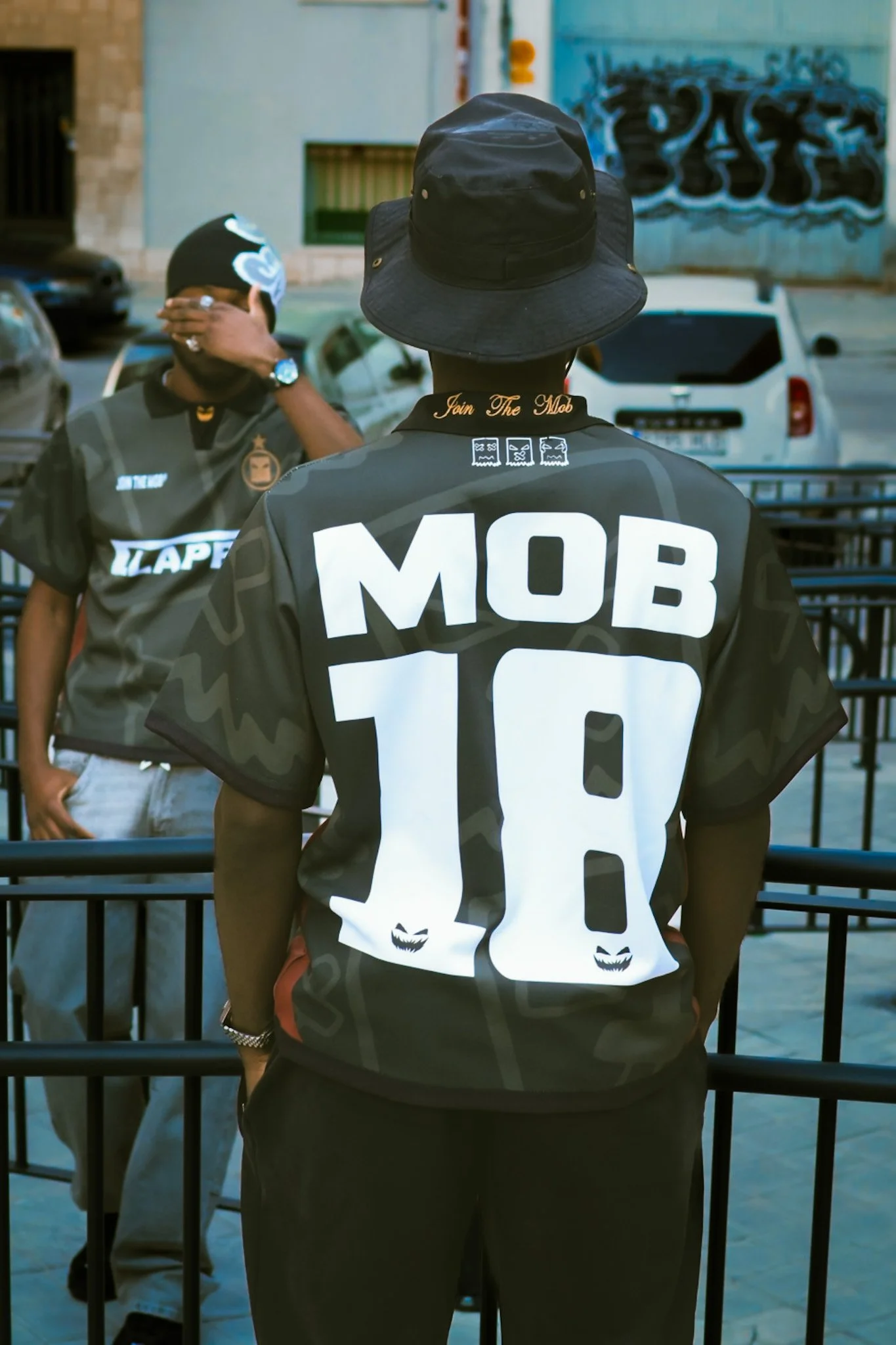 A person wearing a black bucket hat and a black jersey with the words 'Join The Mob' and the number 10 on the back, standing in front of a fence. Two other individuals are visible in the background, one wearing sunglasses and a bandana, and they appe
