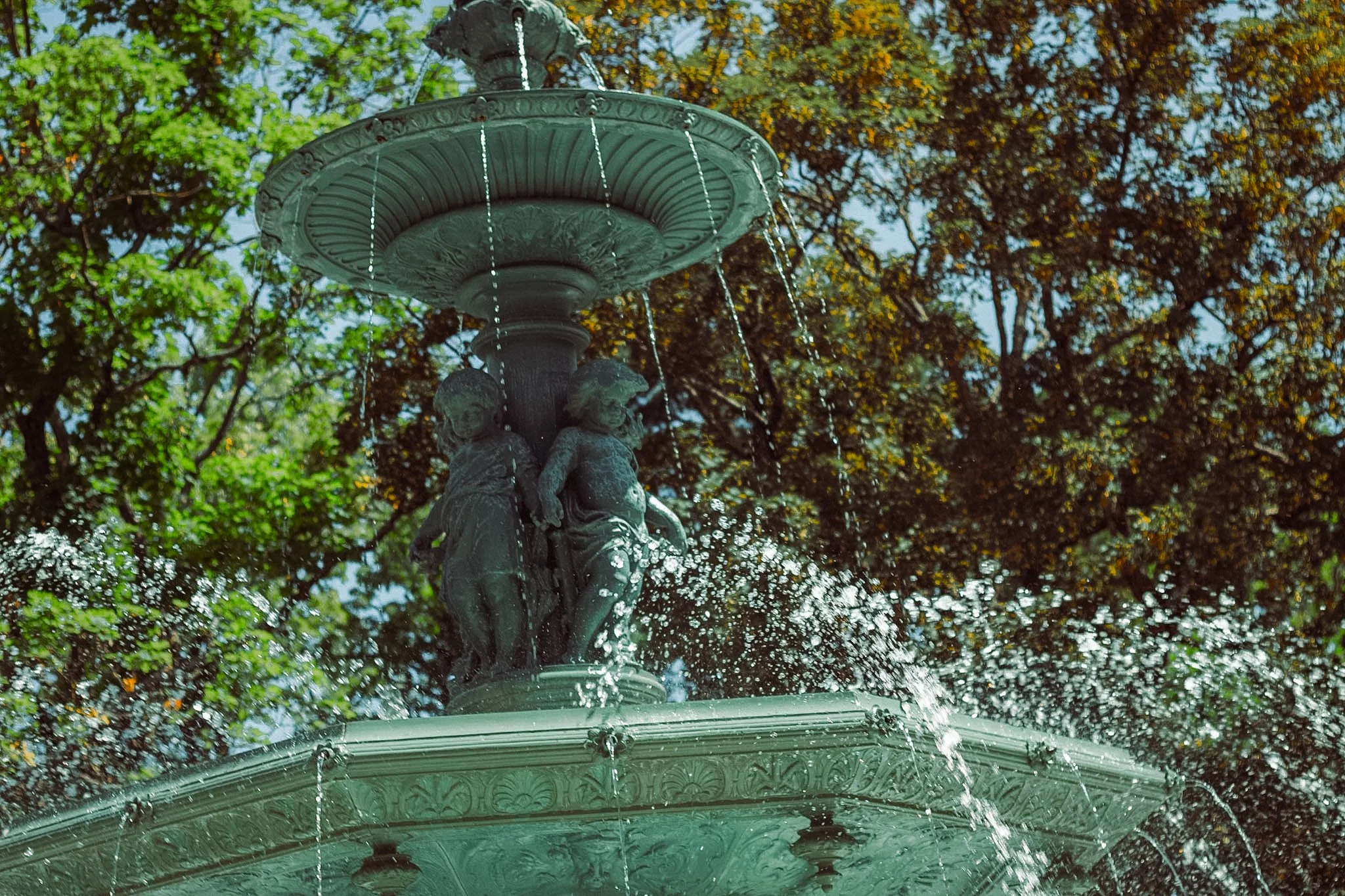 A decorative fountain with statues of children in a park, water flowing from the top tier, surrounded by trees with green and yellow leaves.