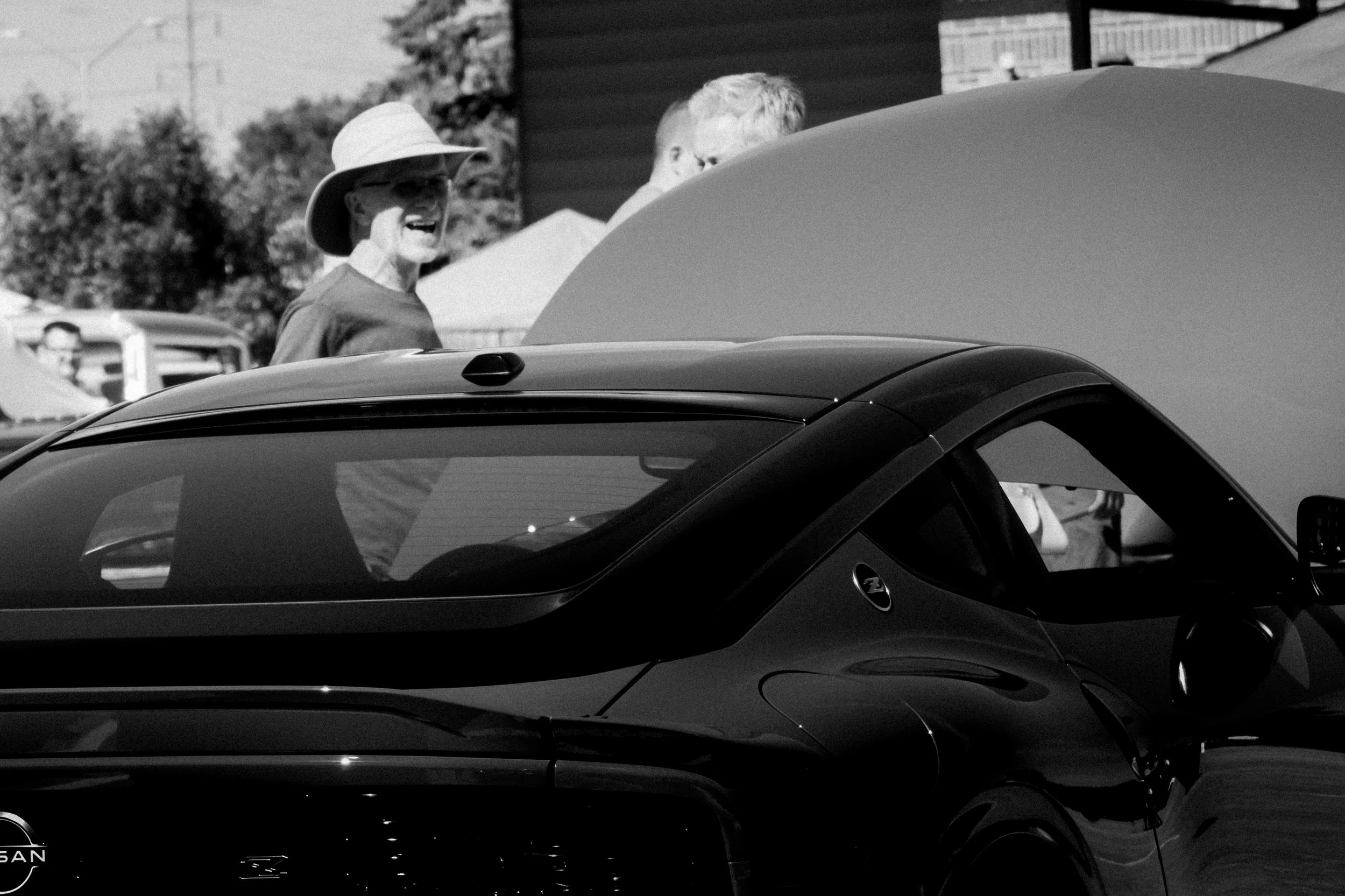 People talking beside a car in an outdoor setting with trees and buildings in the background.