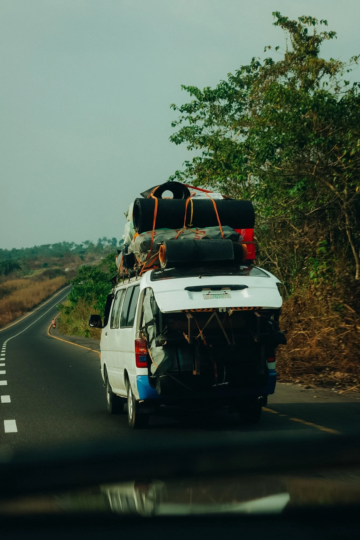 A white van driving on a curvy two-lane road with a load of camping gear and luggage strapped on its roof. The road is surrounded by green trees and a dry field, with a person riding a bicycle on the side of the road.
