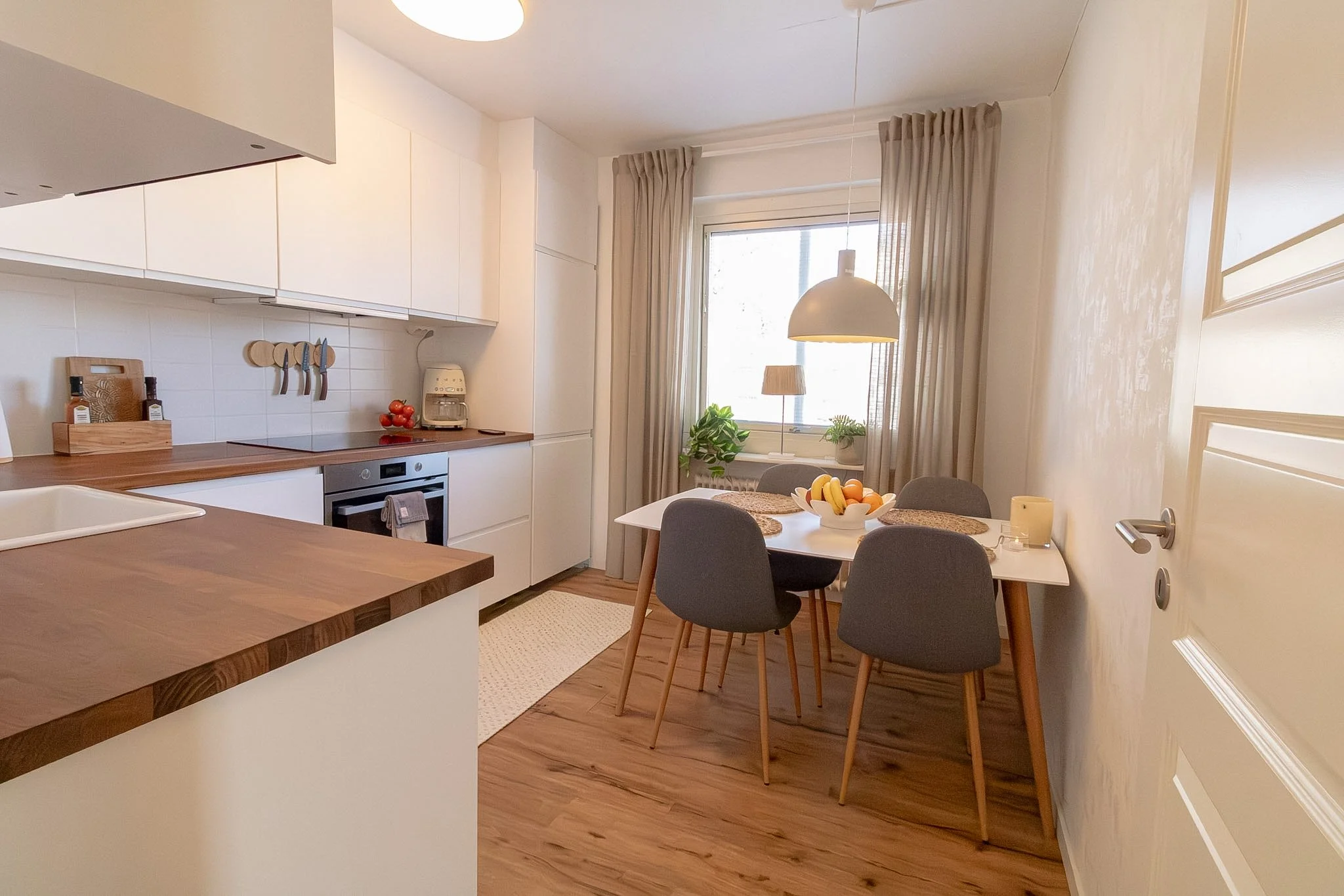 A cozy kitchen and dining area with white cabinets, a round table with four gray chairs, a bowl of fruit, large window with beige curtains, and warm lighting.