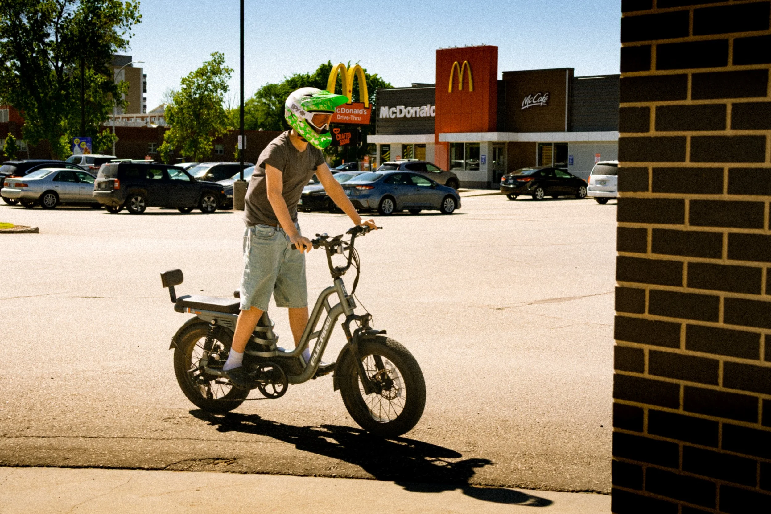 A young person wearing a green helmet with white and purple design, sitting on an electric bike in a parking lot near a brick wall, with a McDonald's and McCafé in the background and trees visible under clear blue sky.