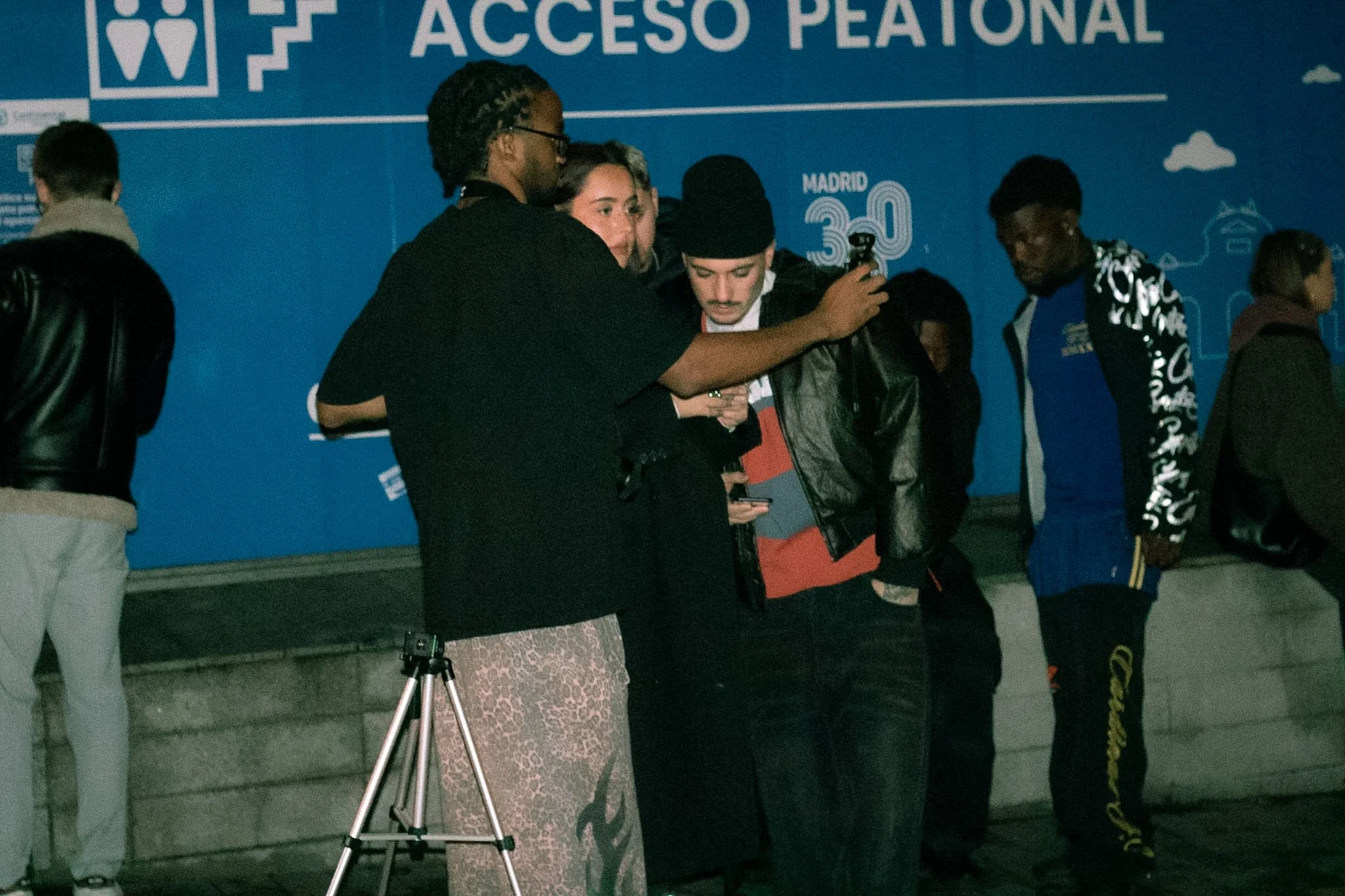 Group of people standing near a blue wall with the words 'ACCESO PEATONAL' and a stair icon, one person taking a photo or video with a smartphone, others engaged or standing around.
