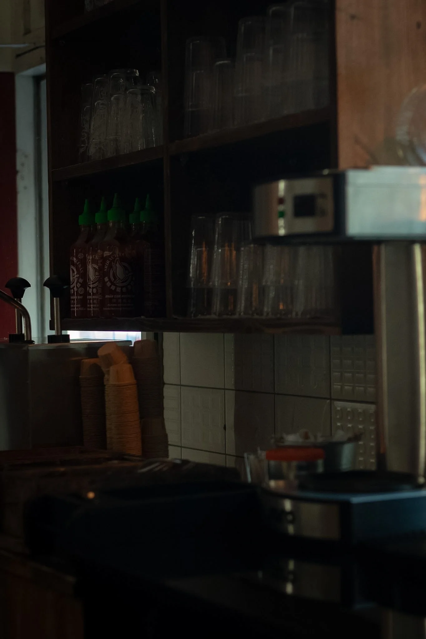 A dimly lit kitchen scene with shelves containing glass bottles, glasses, and paper cups, along with some kitchen appliances and items on the counter.