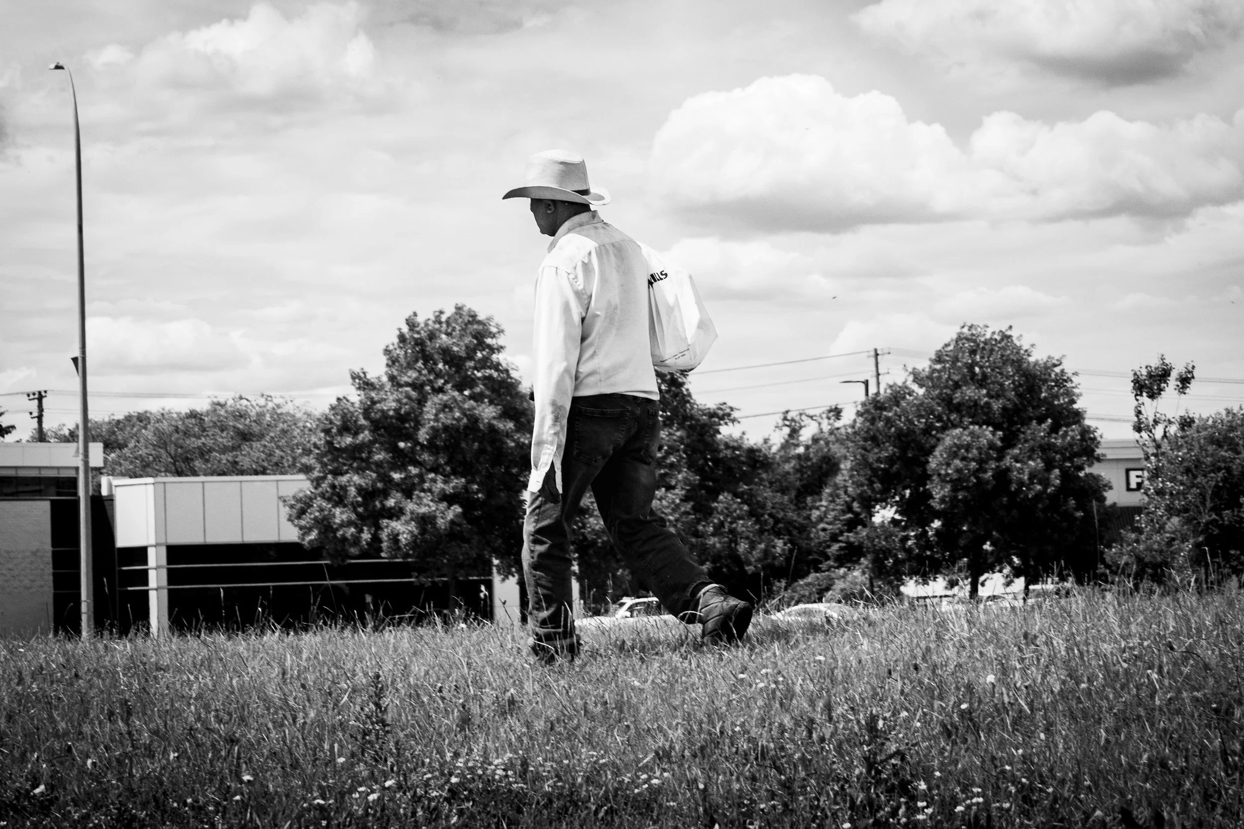 A man in a cowboy hat and jeans walking through grass in an open field with trees, power lines, and clouds in the background, captured in black and white.