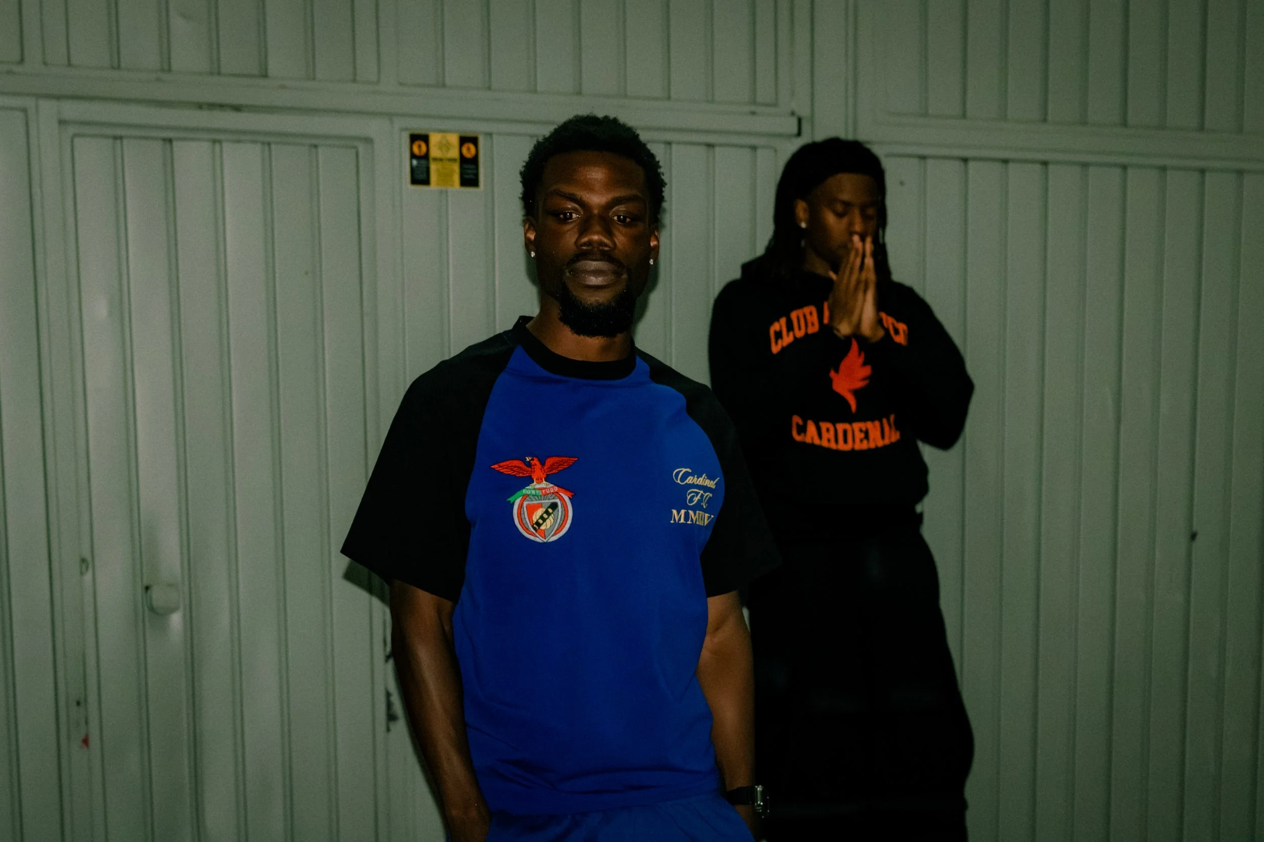Two young men standing against a metal wall inside a building. The man in front is wearing a blue sports jersey with a logo on the chest and looking at the camera. The man in the background has long dreadlocks, is wearing a black hoodie with red and 