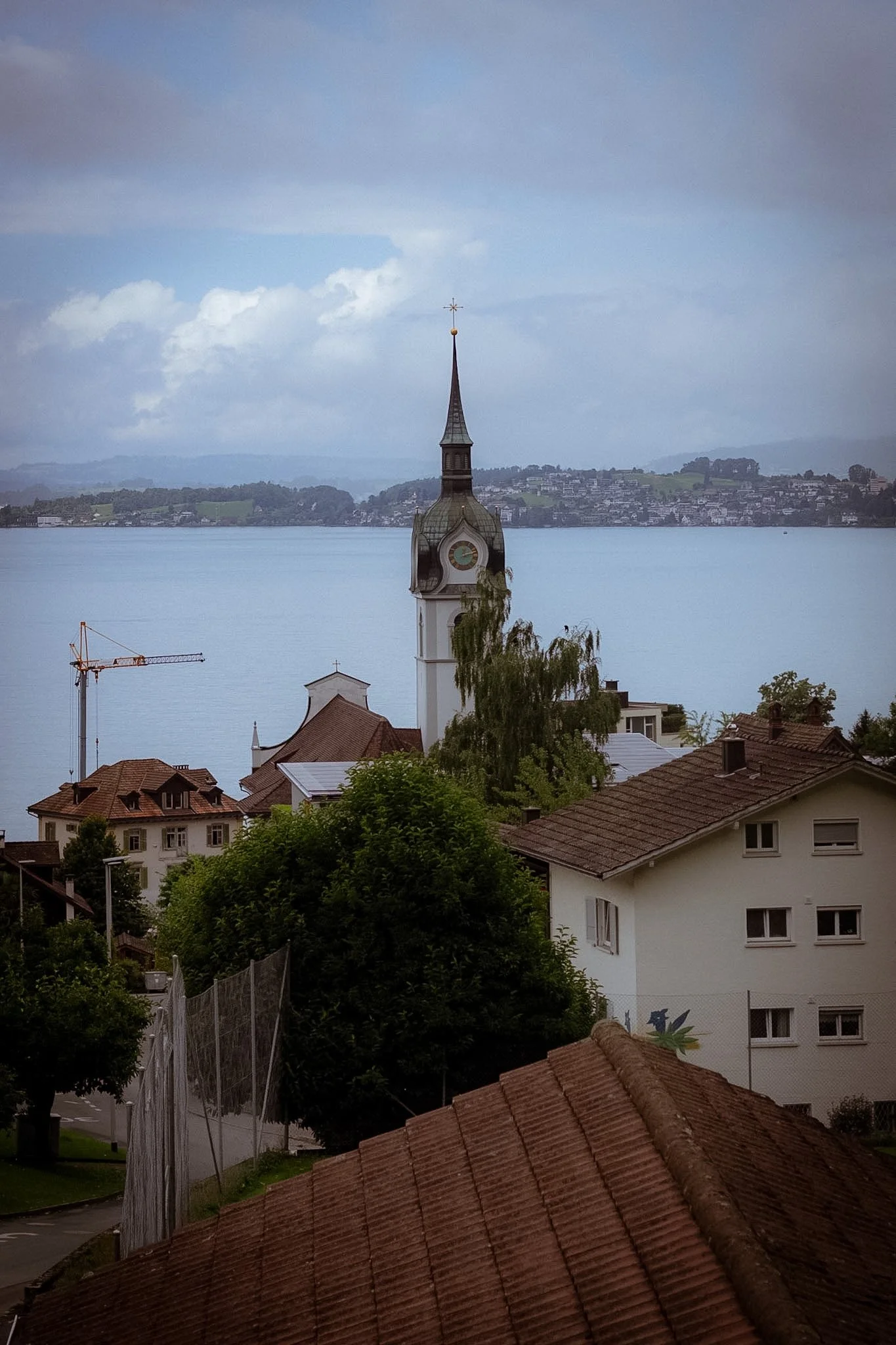 View of a church steeple over residential buildings with red-tiled roofs, trees, and a view of a large body of water in the background under partly cloudy sky.
