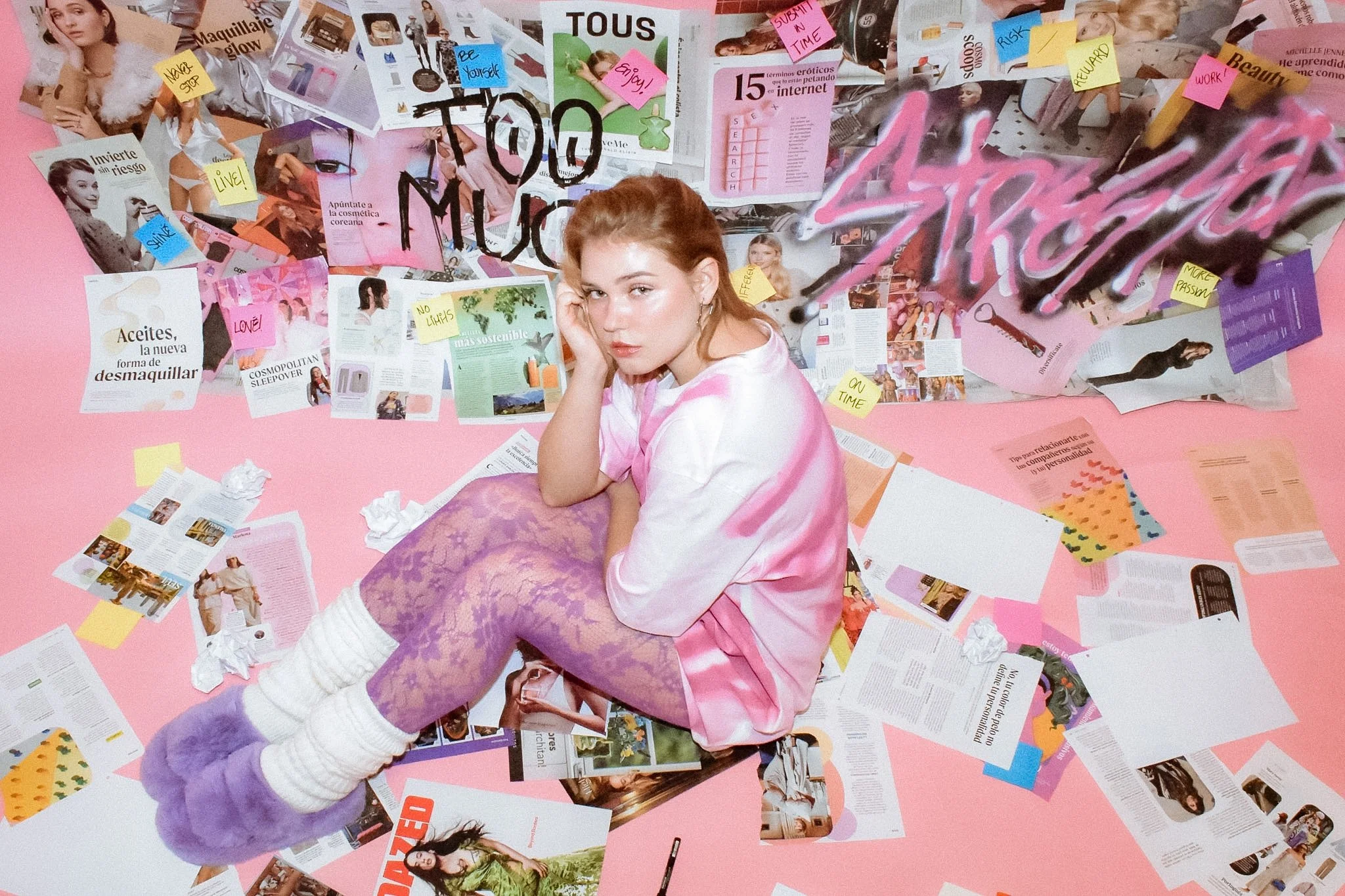A young woman with red hair sits on a pink floor surrounded by scattered magazines, papers, and sticky notes, looking directly at the camera with a contemplative expression.