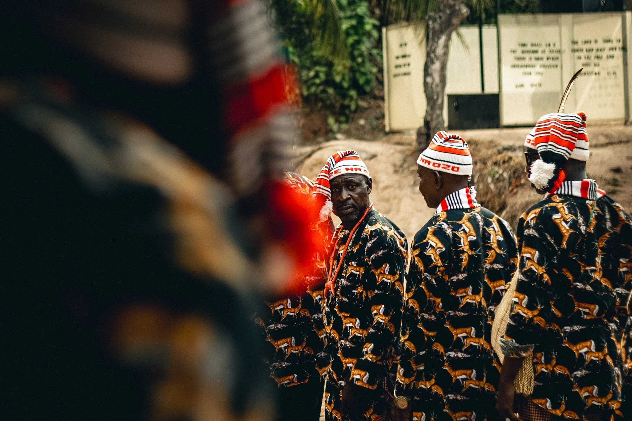 Group of people wearing matching African print outfits and striped hats, standing outdoors near trees and informational signs.
