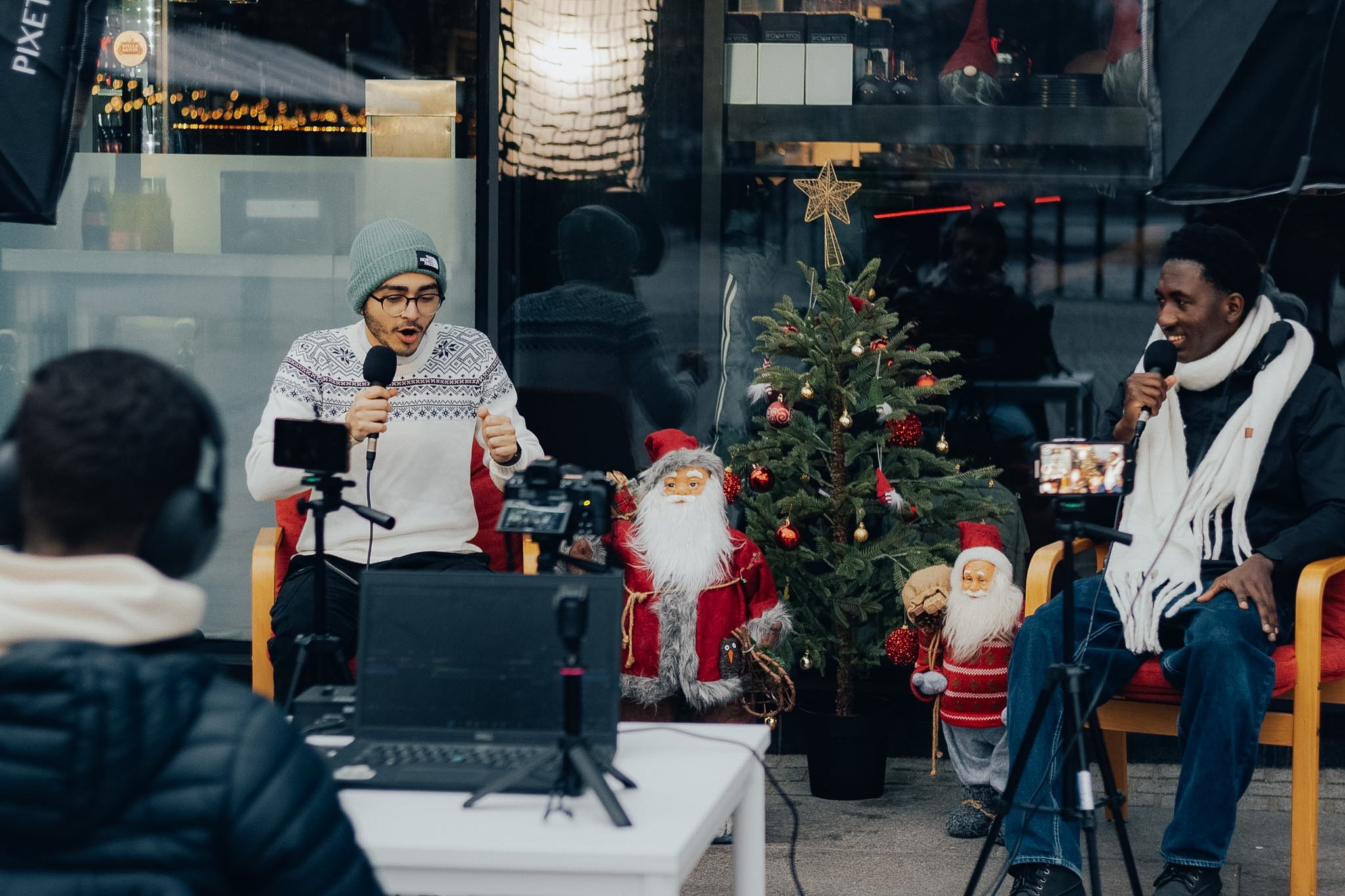 Two men sitting in front of a Christmas tree with ornaments and figurines, engaging in a conversation or interview, with cameras capturing the scene.