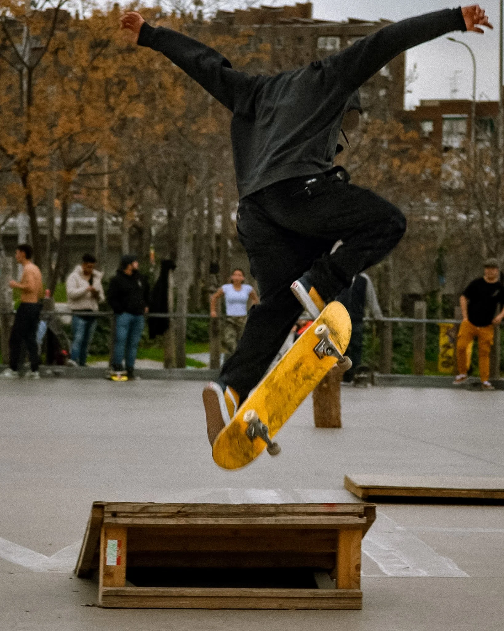 A person skateboarding off a wooden ramp in an outdoor skate park during the evening, with several onlookers in the background and trees with autumn foliage.
