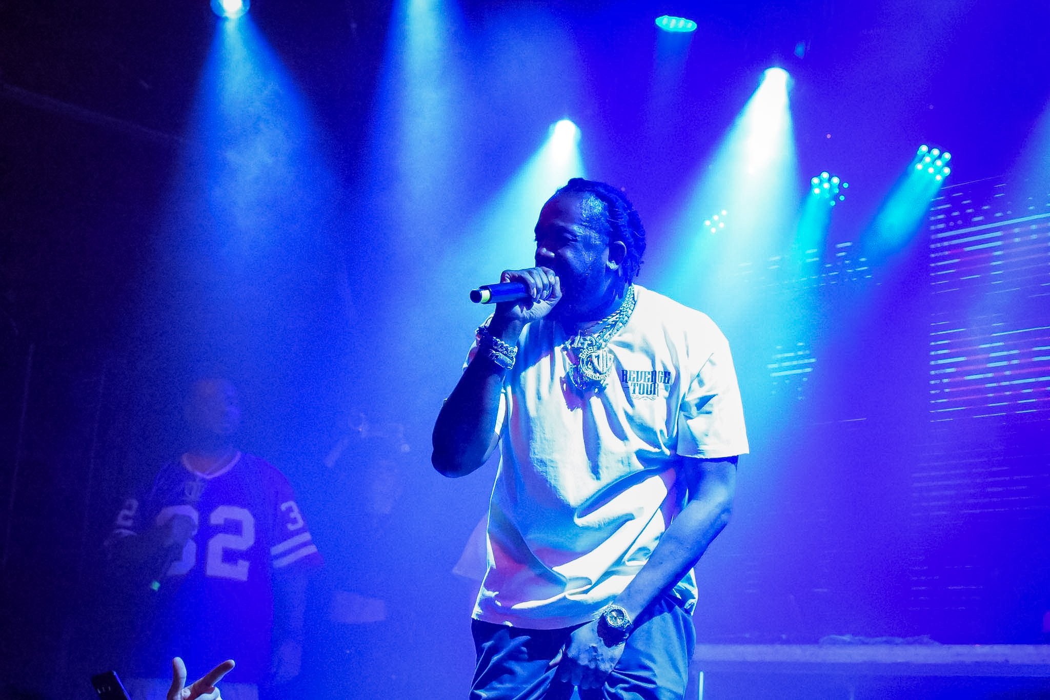 A male singer performing on stage under blue lights, wearing a white t-shirt and jewelry, holding a microphone close to his mouth.