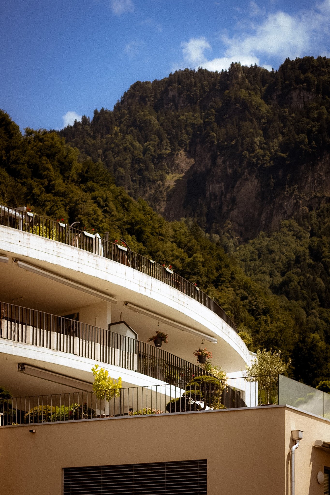 Multiple levels of a modern white building with black railings and outdoor plants, set against a mountainous background with a blue sky and some clouds.