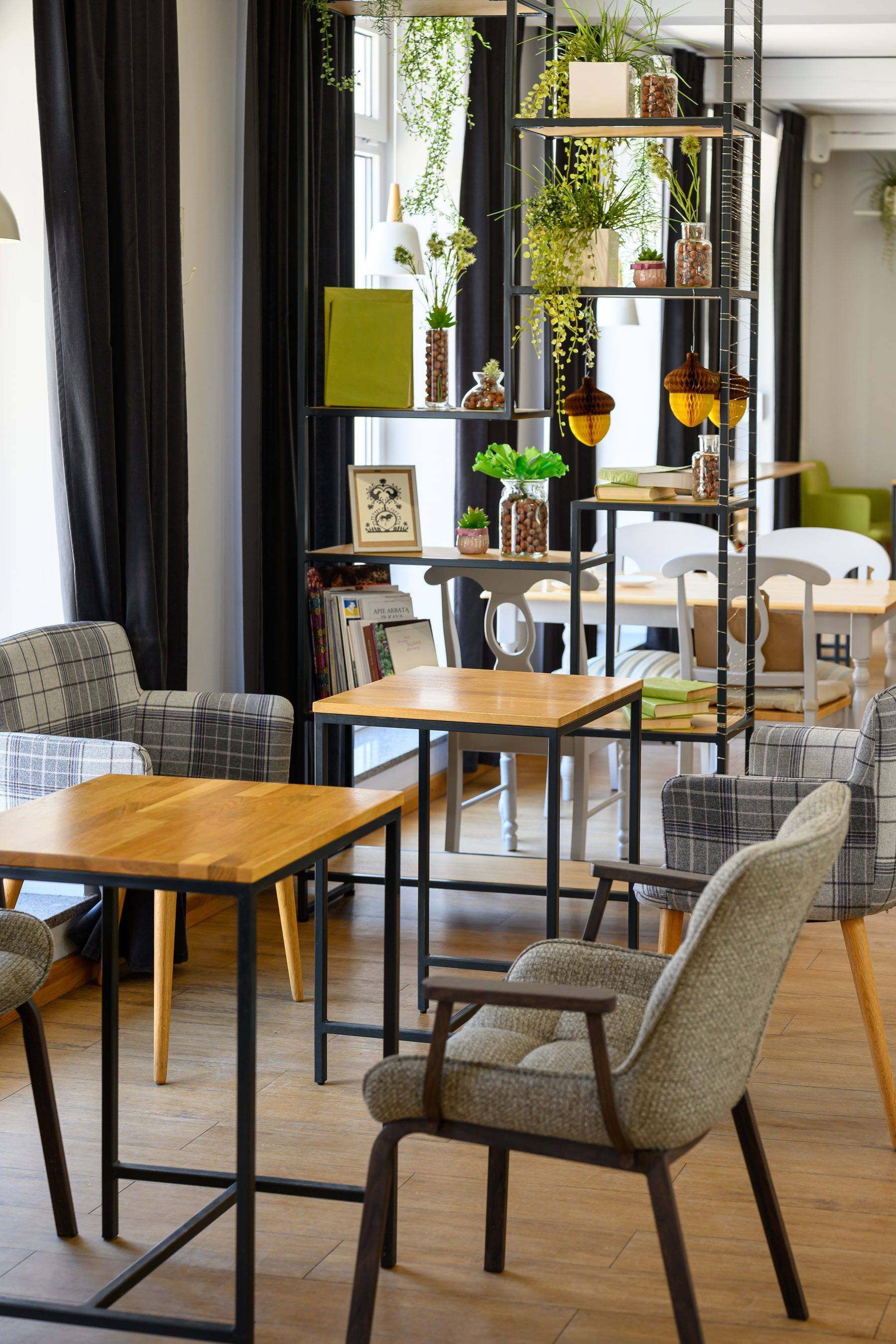 A cozy modern dining and lounge area with wooden tables and upholstered chairs, separated by a black metal shelving unit decorated with green plants, books, and glass jars filled with snacks, near large windows with black curtains.