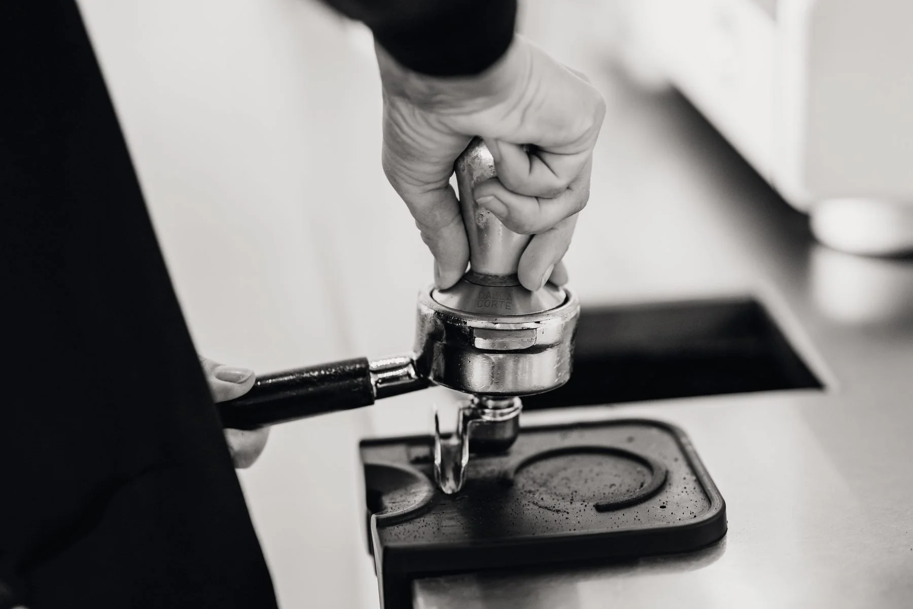 A person with a glove is using a tamper to compress coffee grounds in a portafilter on a stovetop espresso machine.