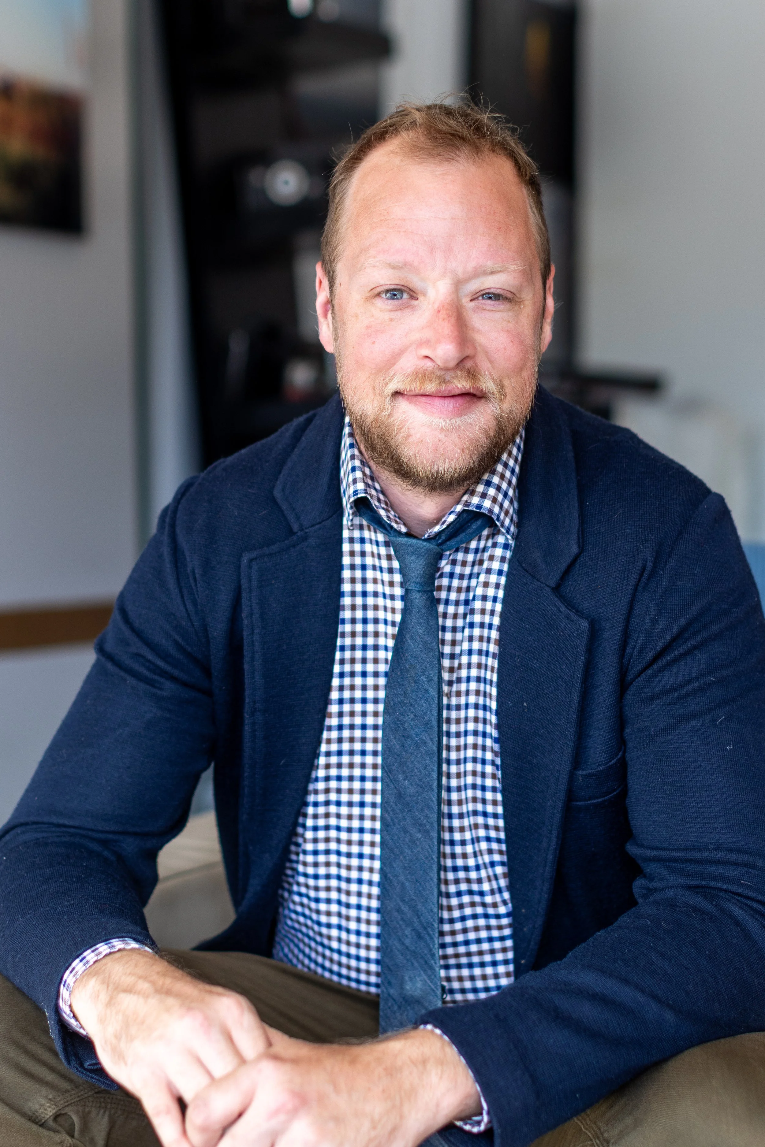 A man with red hair and a beard smiling, sitting at a desk with his arms crossed, wearing a checkered shirt, blue blazer, and gray tie in an office setting.
