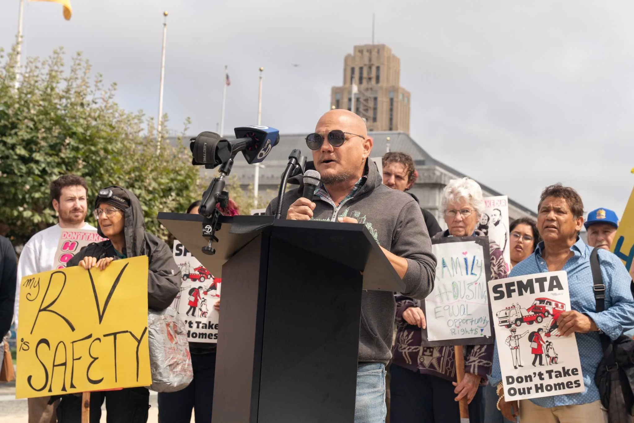 A man (SDA Housing Organizer Ocean Coast) stands at a podium speaking into a microphone. Behind him, people are holding signs that say: “RV Safety”, “SFMTA Don’t Take Our Homes”, and “Family Housing Equal Opportunity Rights”.