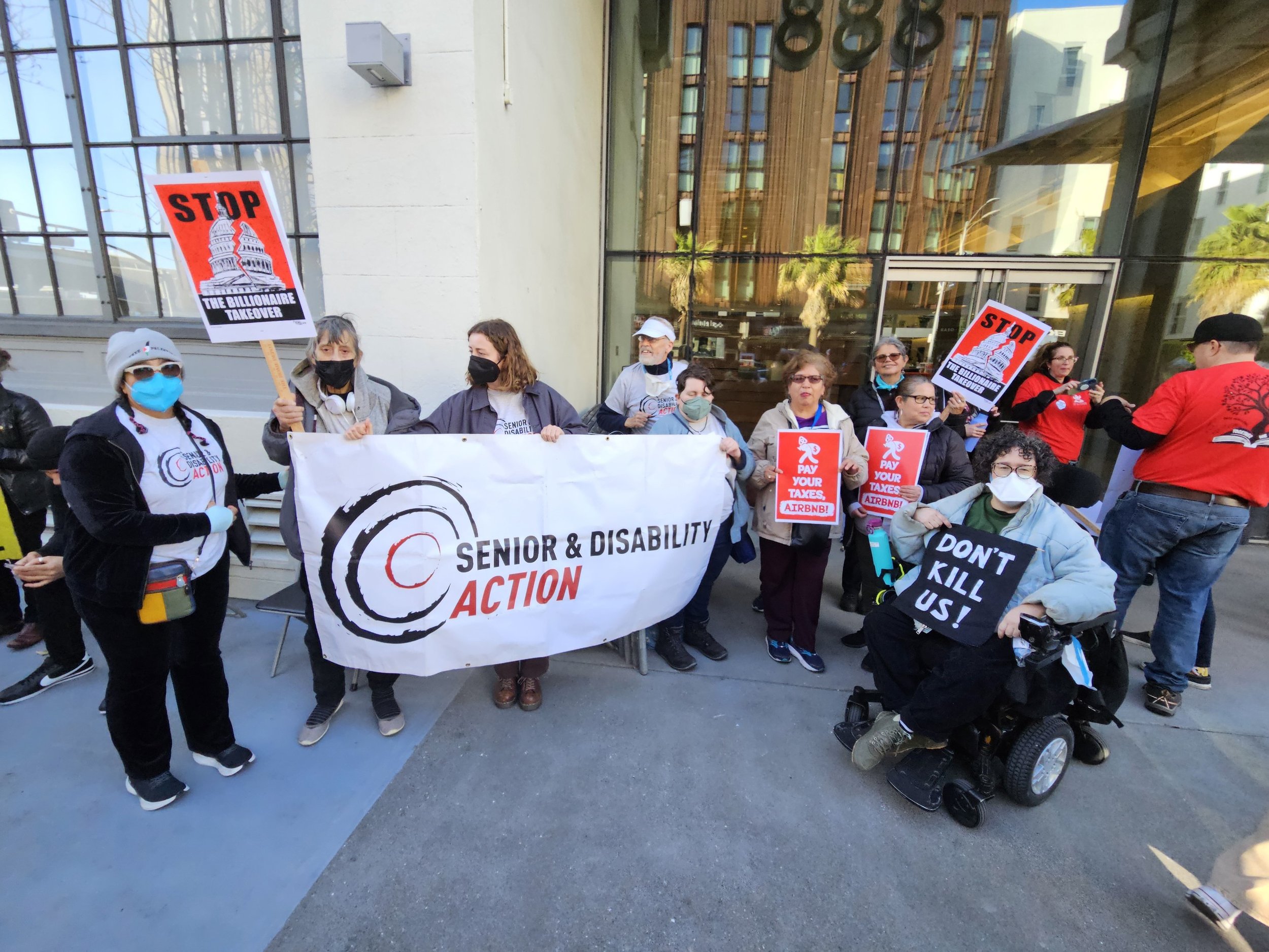 A group of masked seniors and people with disabilities holding signs that say “Stop the billionaire takeover” and “pay your taxes, Airbnb” in front of the SDA banner.