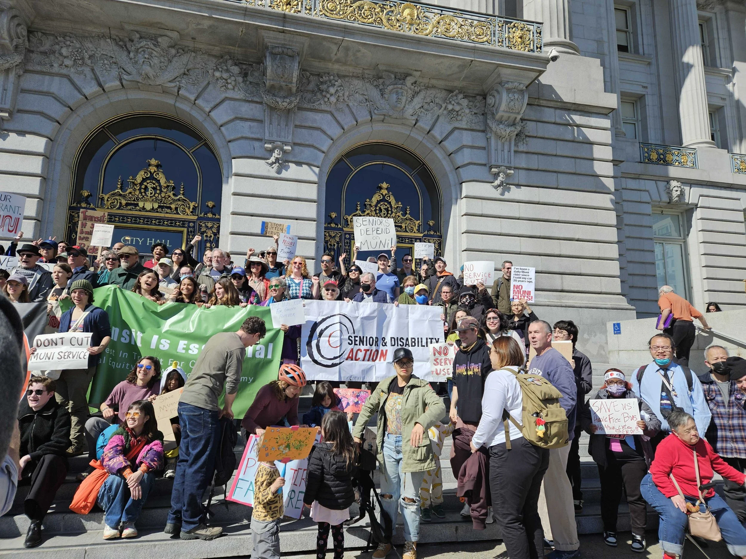 A large group of people on the steps of City Hall, holding signs that say Don’t Cut Muni Service, and banners including SDA's