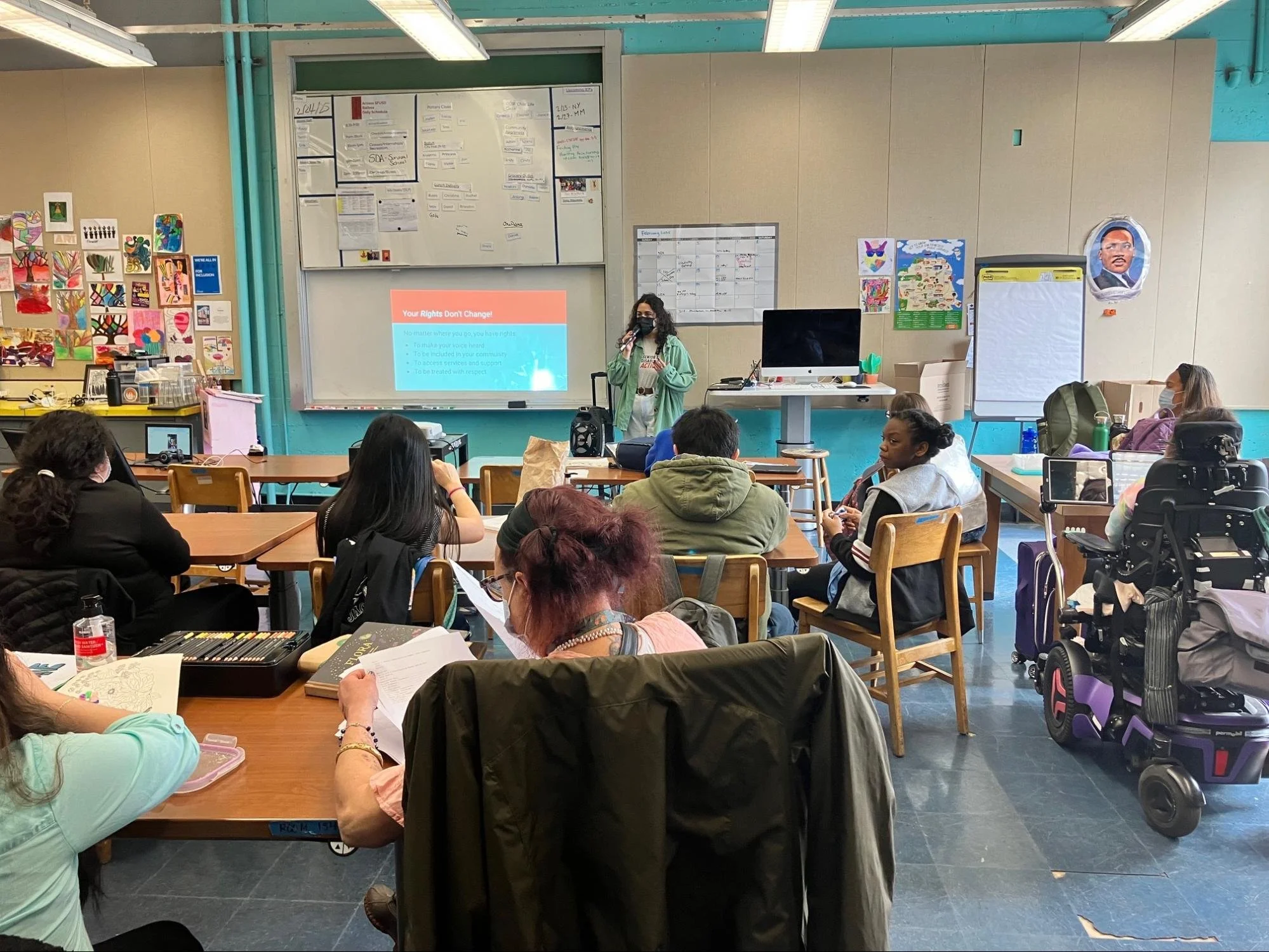 Students sitting at tables in a classroom, as someone in a mask stands at the front of the room by a projector screen.