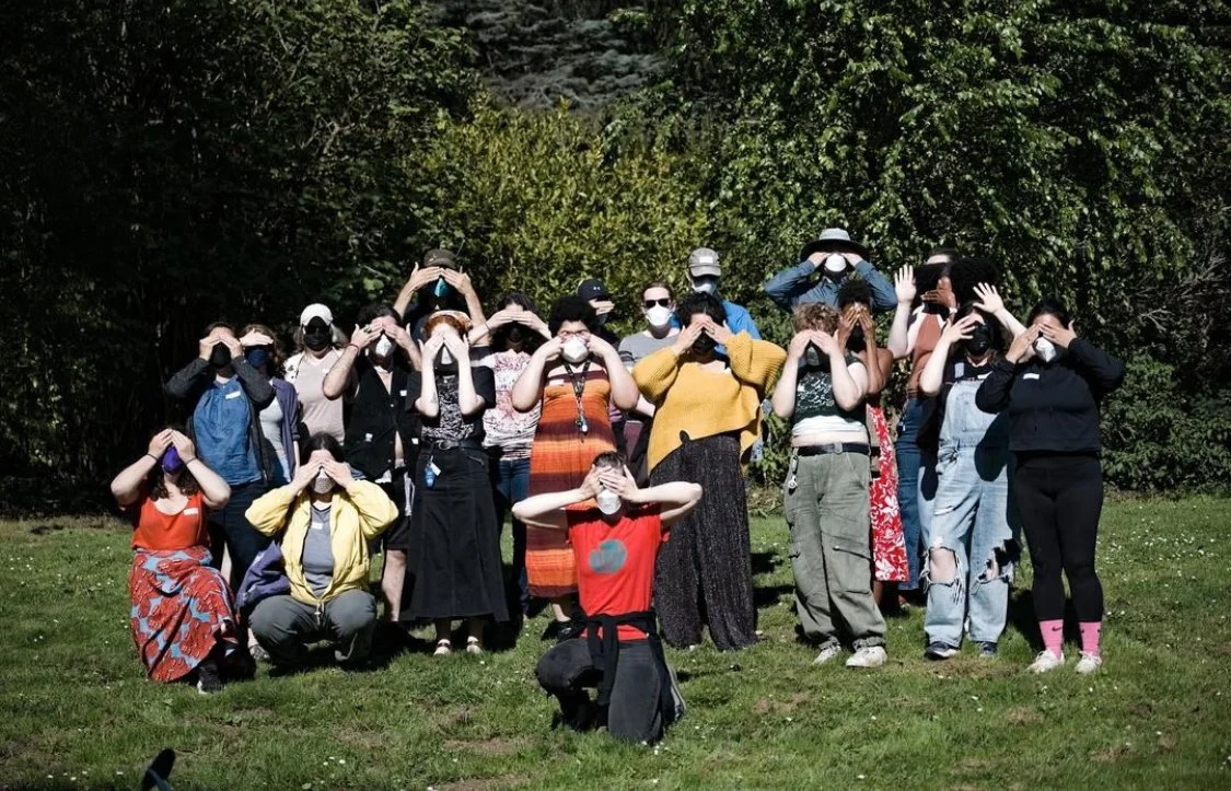 22 personas posan juntas para una foto con máscaras en el Golden Gate Park. Se cubren los ojos con las manos y se paran sobre la hierba verde con arbustos y árboles detrás de ellos. Tres personas están arrodilladas y el resto del grupo de pie.