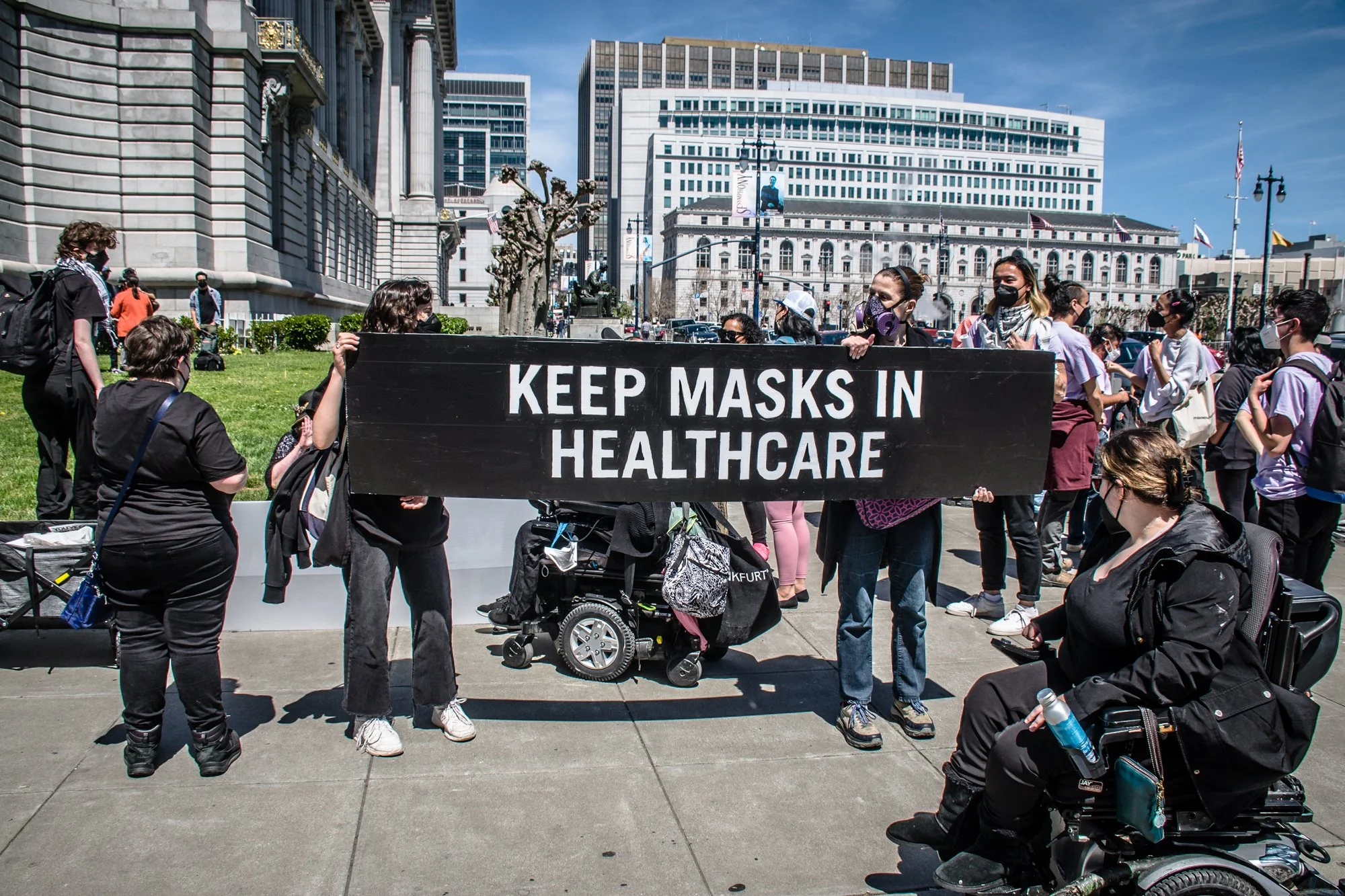Un gran grupo de personas enmascaradas afuera en una esquina, incluidas dos en primer plano que sostienen un cartel que dice "mantenga las máscaras en la atención médica".