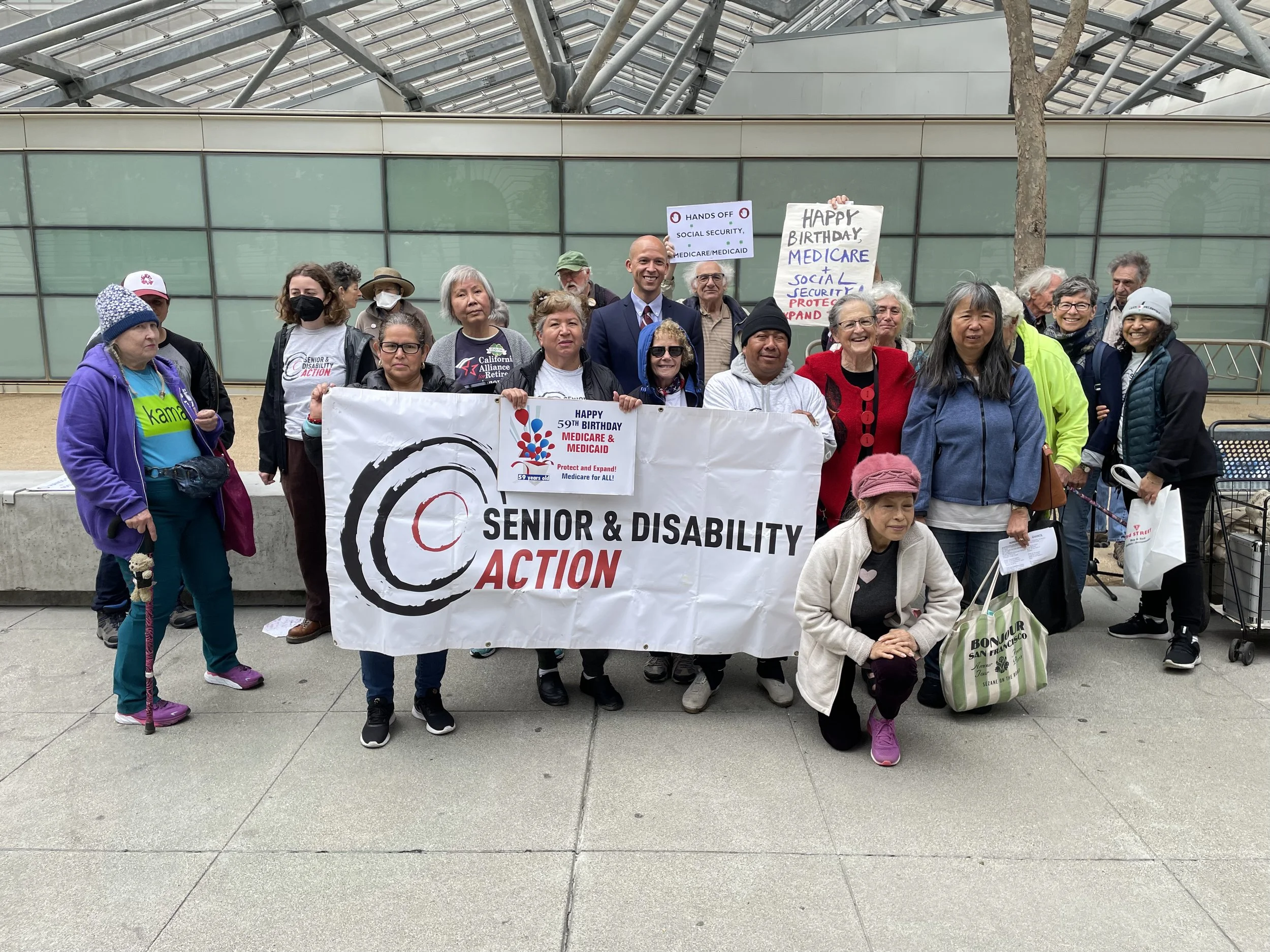 A group of people stands in front of a banner that says Senior and Disability Action. Others hold smaller signs that say Happy 59th Birthday Medicare and Medicaid and Happy Birthday Medicare & Social Security.