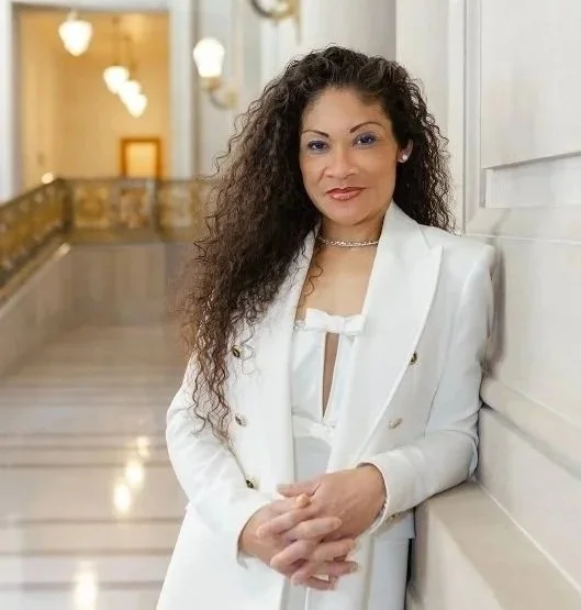 A woman with long curly hair wearing a white suit and leaning against the wall in a building with a marble floor