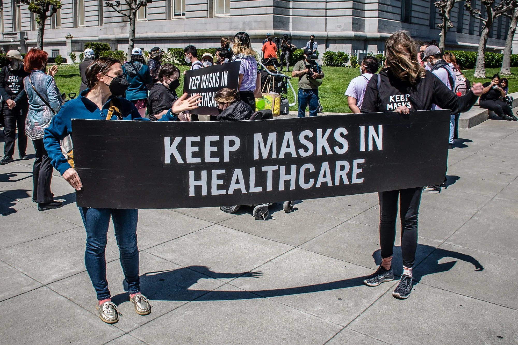 Un gran grupo de personas enmascaradas afuera en una esquina, incluidas dos en primer plano que sostienen un cartel que dice "mantenga las máscaras en la atención médica".