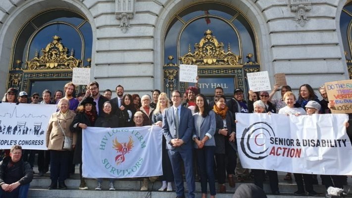 Un grupo de aproximadamente 40 personas de pie en las escaleras del Ayuntamiento. Llevan pancartas que dicen "Congreso del Pueblo", "Sobrevivientes del VIH a largo plazo" y "Acción para personas mayores y discapacitadas".
