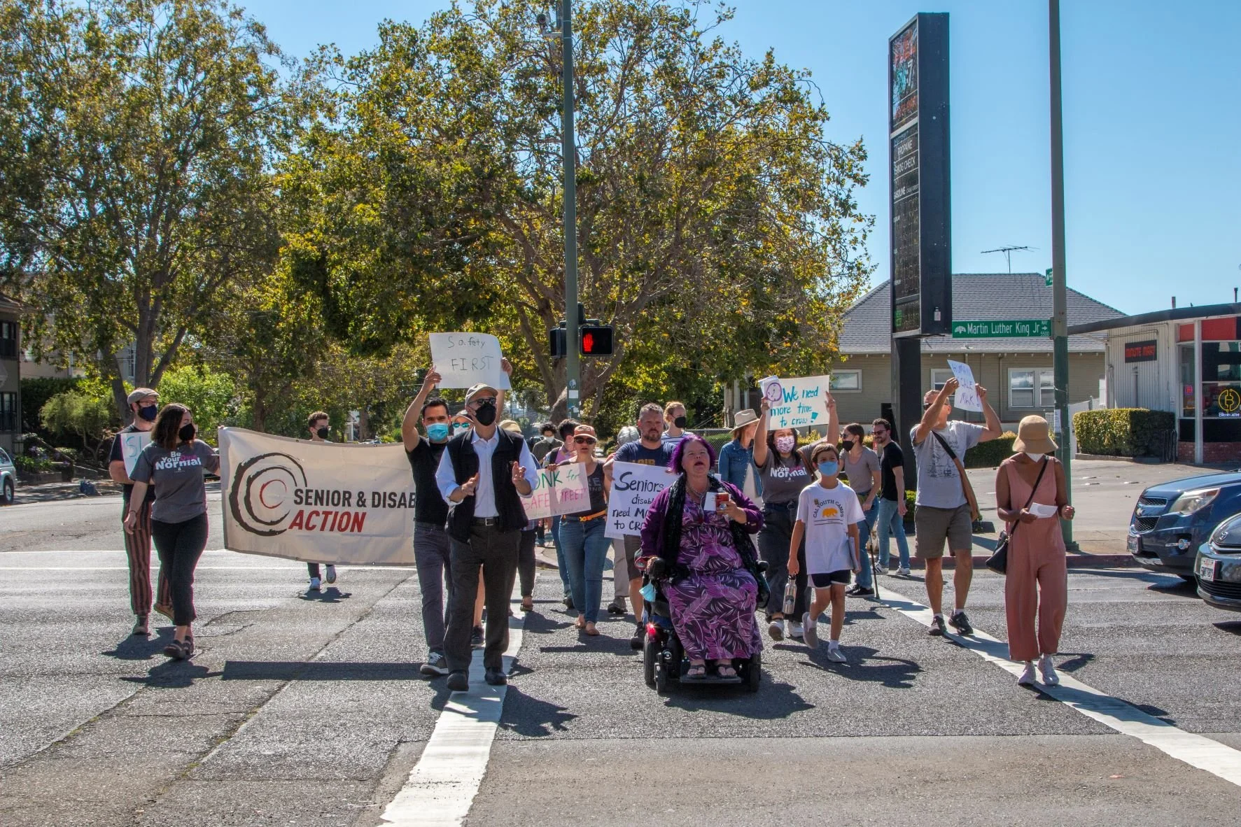 A group of people crossing the street on a red light. One of them is in a wheelchair. They are holding signs that say “Safety first!” and “We need more time to cross.”