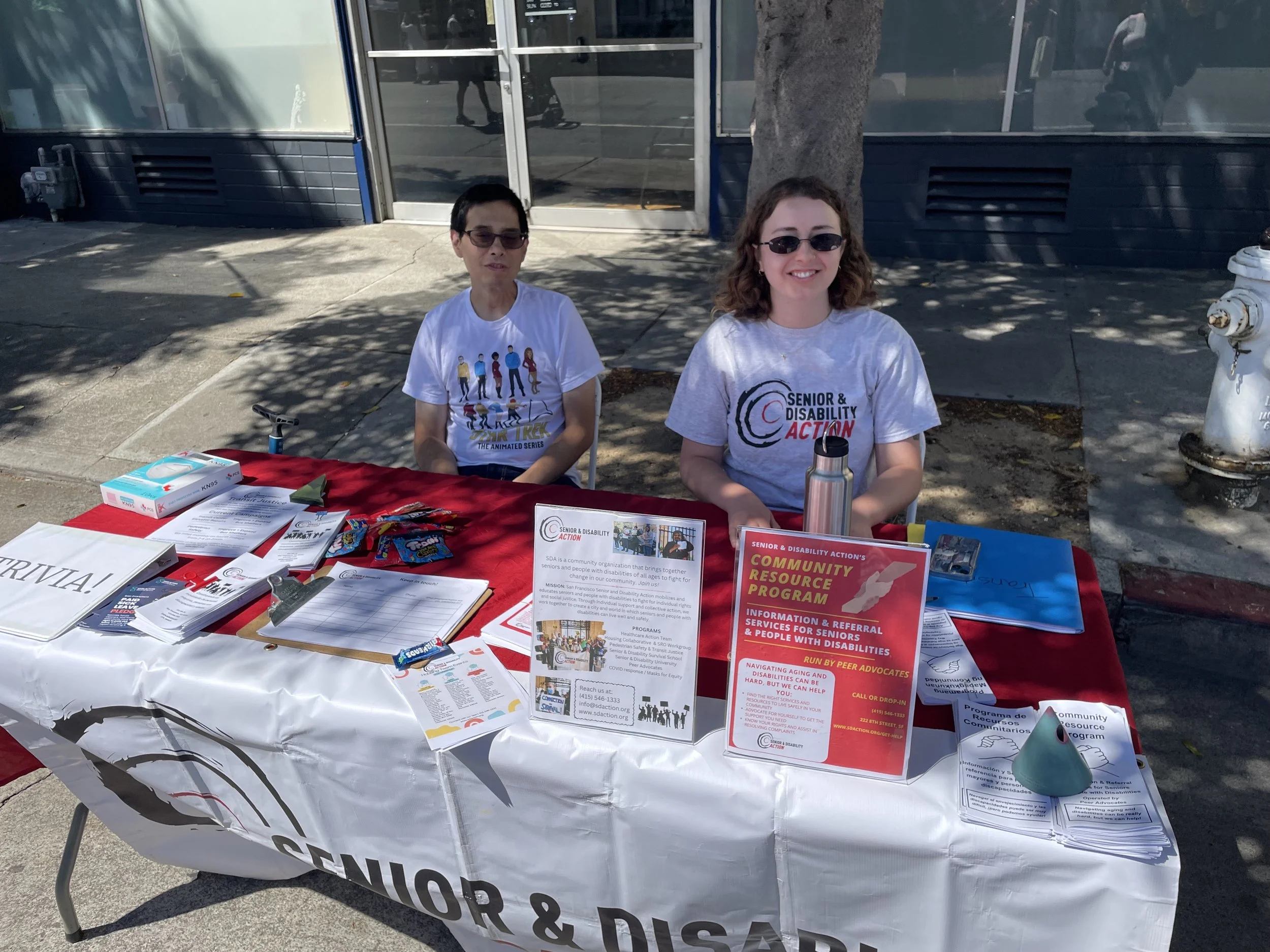 Two people sit outside behind a table with a banner that says Senior and Disability Action.