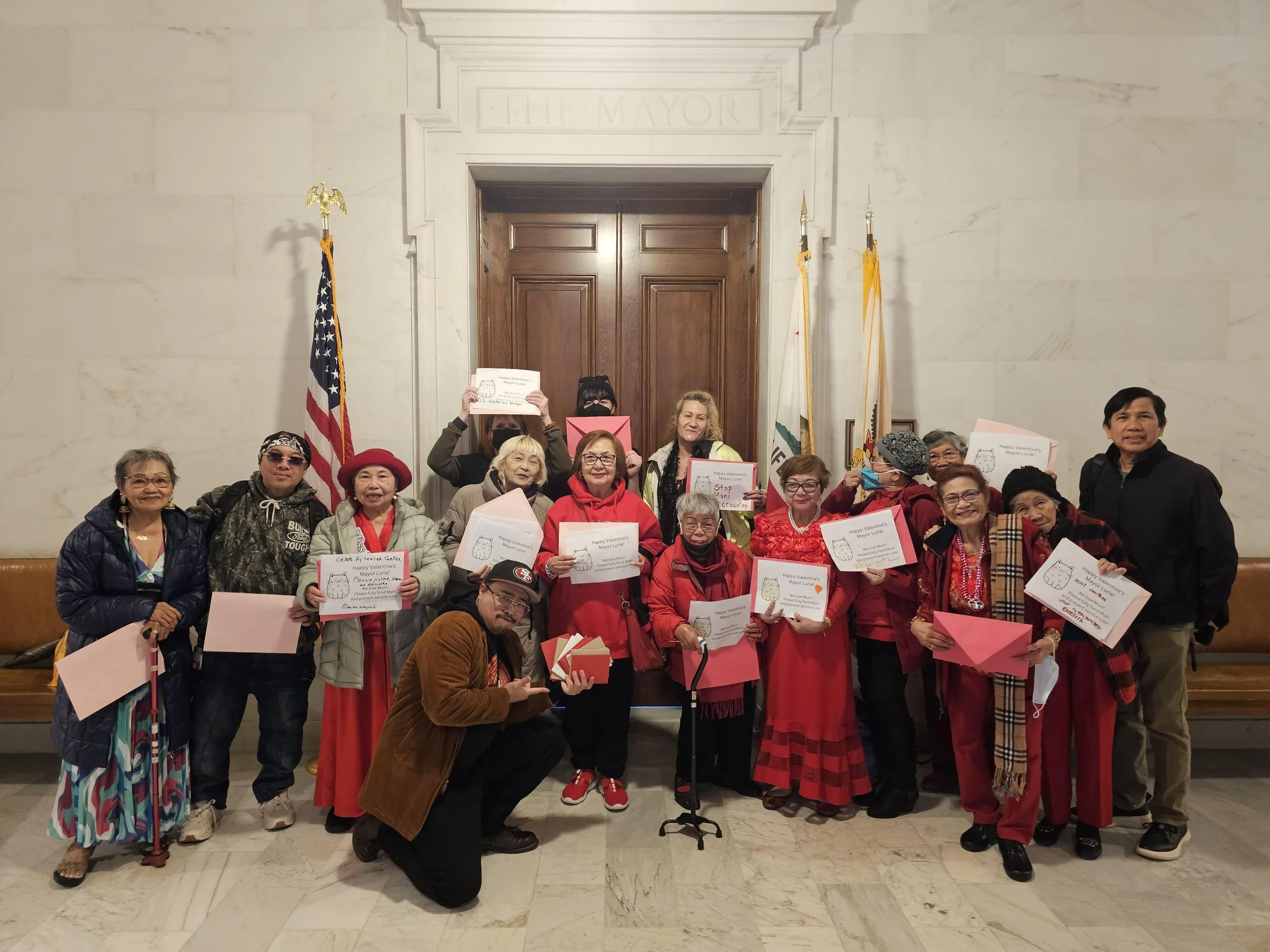 A group of people, many of them seniors dressed in red, holding up valentines in front of a door in a government building