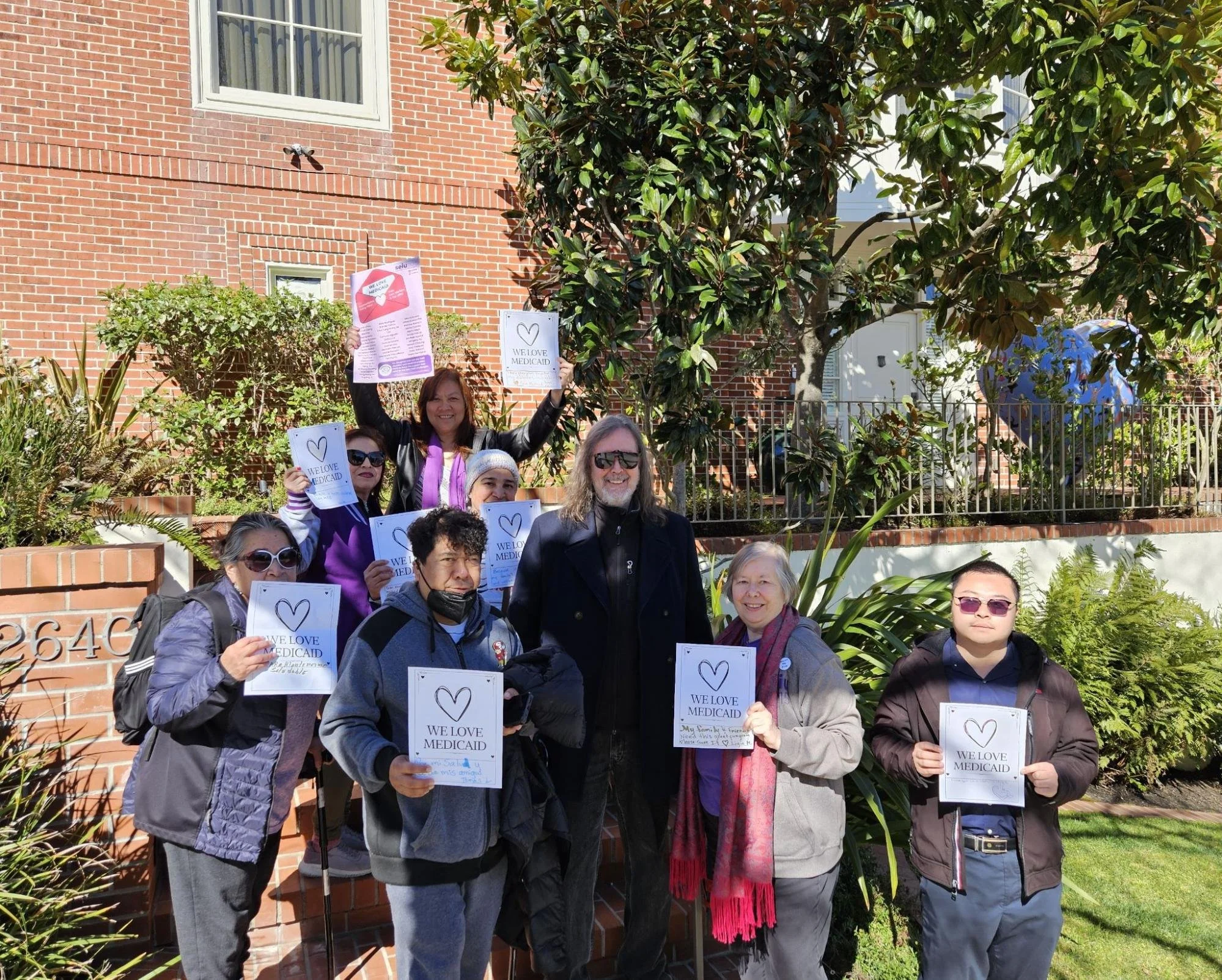 A group of people standing outside a brick house smiling and holding papers that say “We love Medicaid"