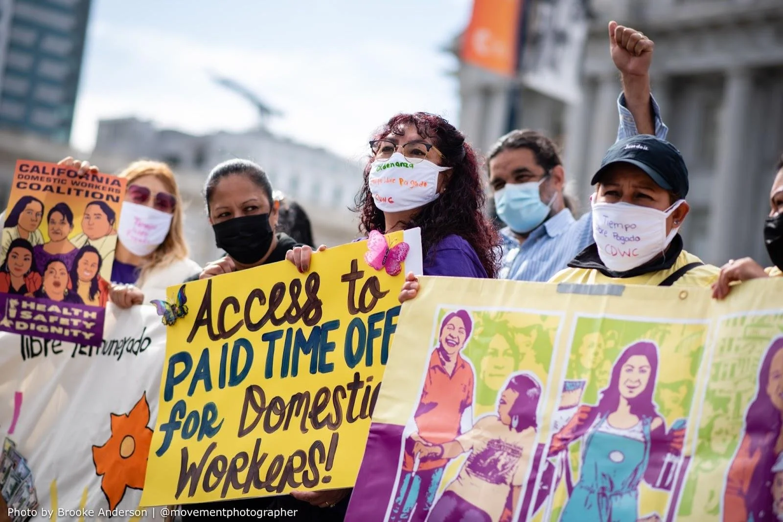 A group of people wearing masks, one with a fist in the air. One holds a sign for the California Domestic Workers Coalition and one holds a sign that says "Access to paid time off for domestic workers!"