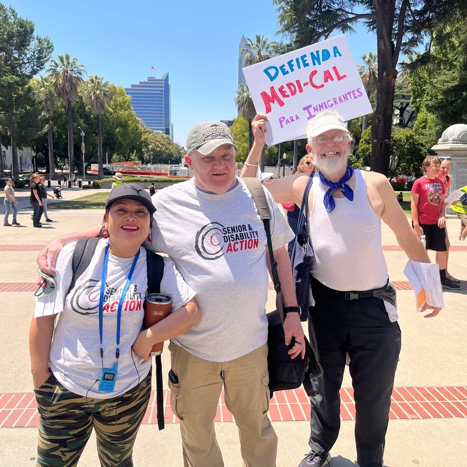 Three people wearing shirts that say Senior and Disability Action and smiling. One of them holds a sign that says “Defienda Medi-Cal para inmigrantes"