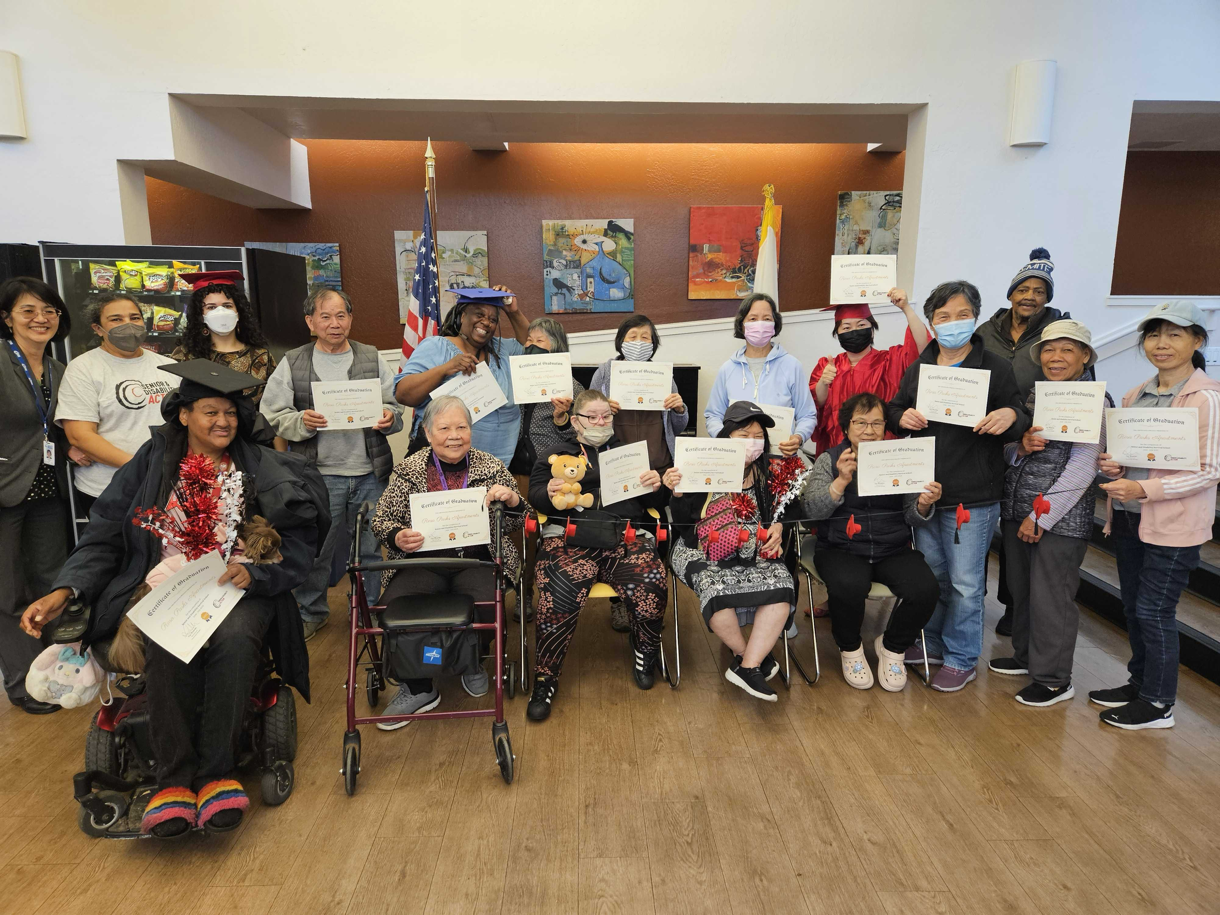 A group of seniors standing and seated in chairs and wheelchairs, holding diplomas