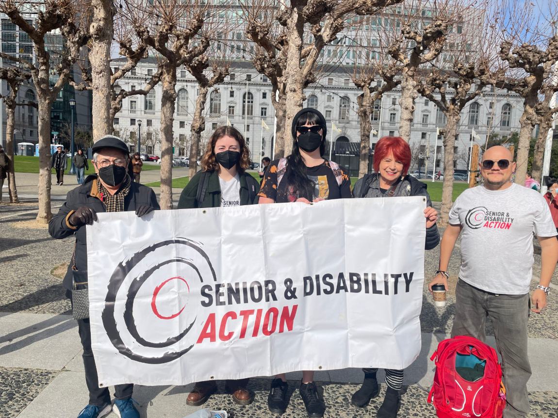 A group of five people, three of them wearing masks, stands outside in front of trees and holding a banner that says Senior and Disability Action