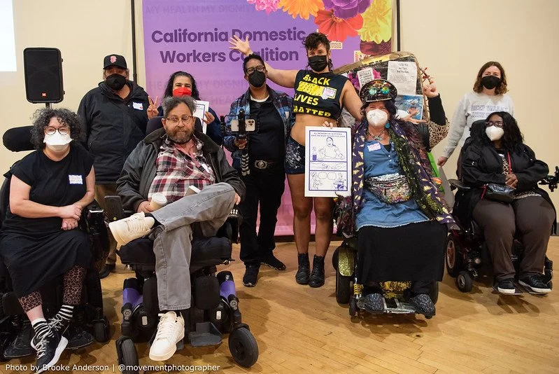 A multiracial group of people standing and sitting in power wheelchairs, in front of a banner that says California Domestic Workers Coalition.