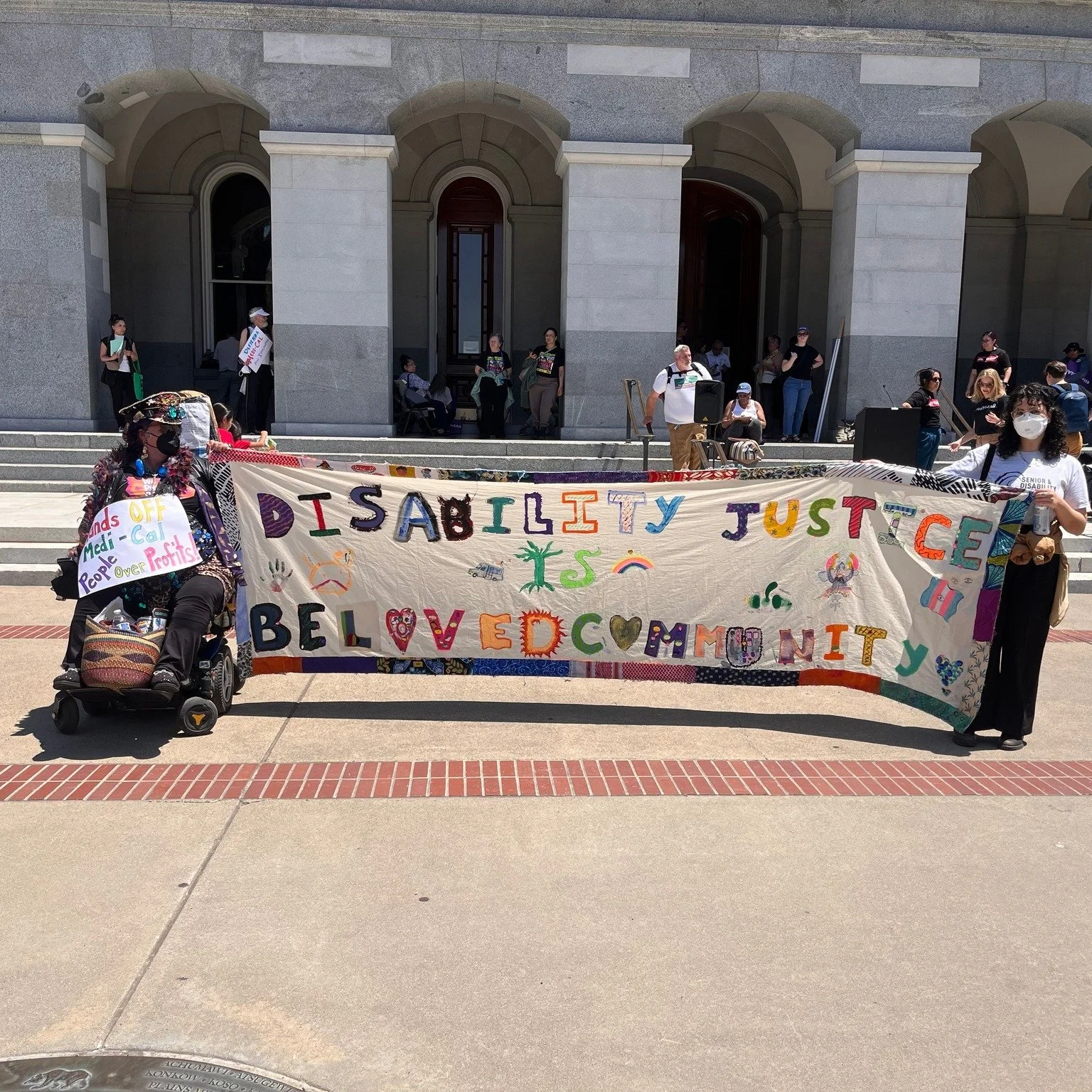 Two people holding a banner that says “Disability justice is beloved community”. They are wearing masks and one of them is in a power wheelchair. There are people standing by a building behind them.