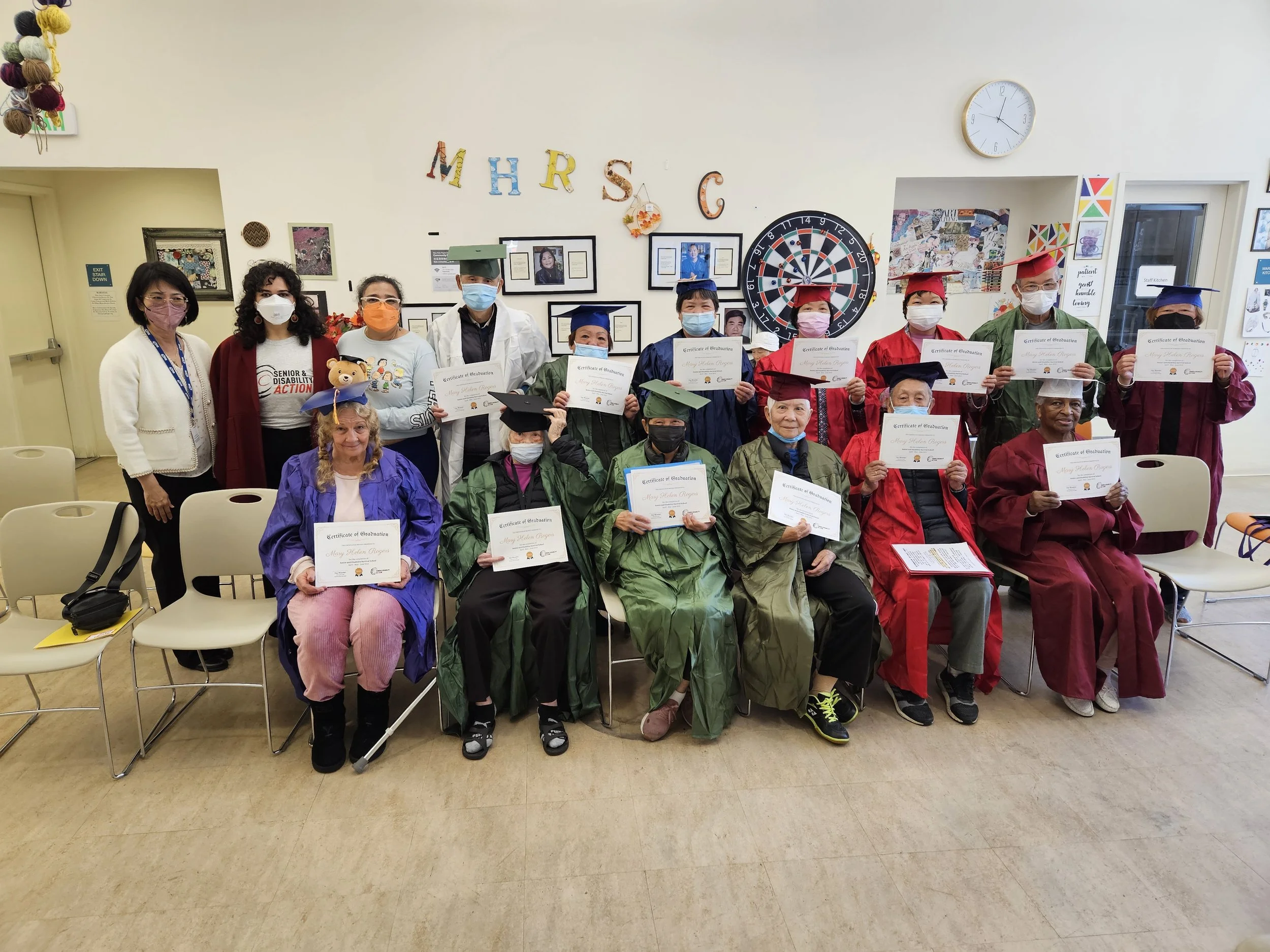 A group of masked people sitting and standing, many of them wearing graduation caps and gowns and holding certificates