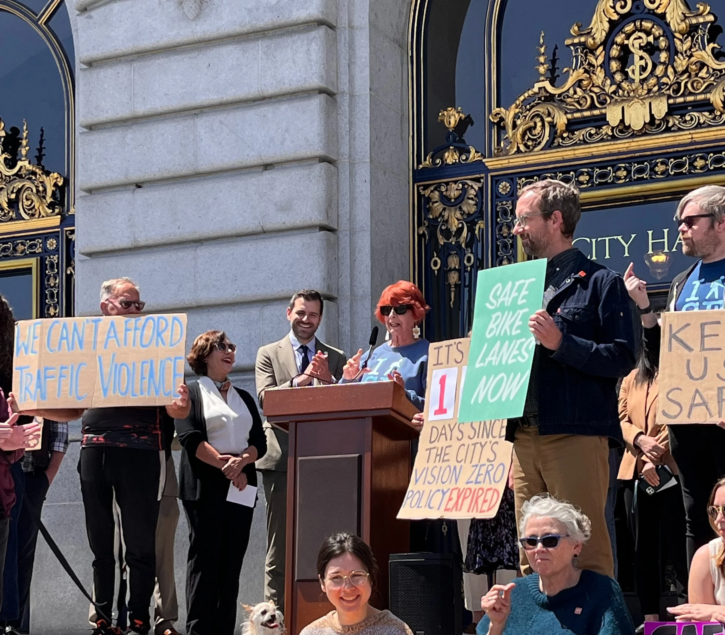 A senior woman with red hair speaks at a podium surrounded by people smiling, including Supervisors Melgar and Sauter. People hold signs that say “we can’t afford traffic violence” and “safe bike lanes now"